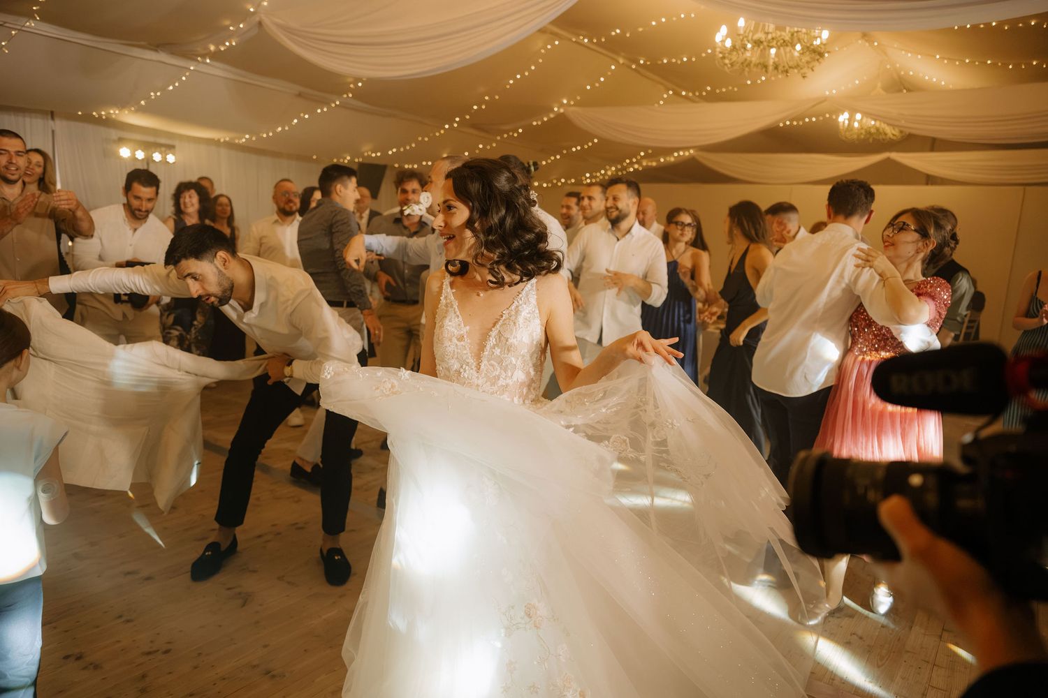 Bride twirling in white gown on wedding reception dance floor surrounded by guests and photographer.