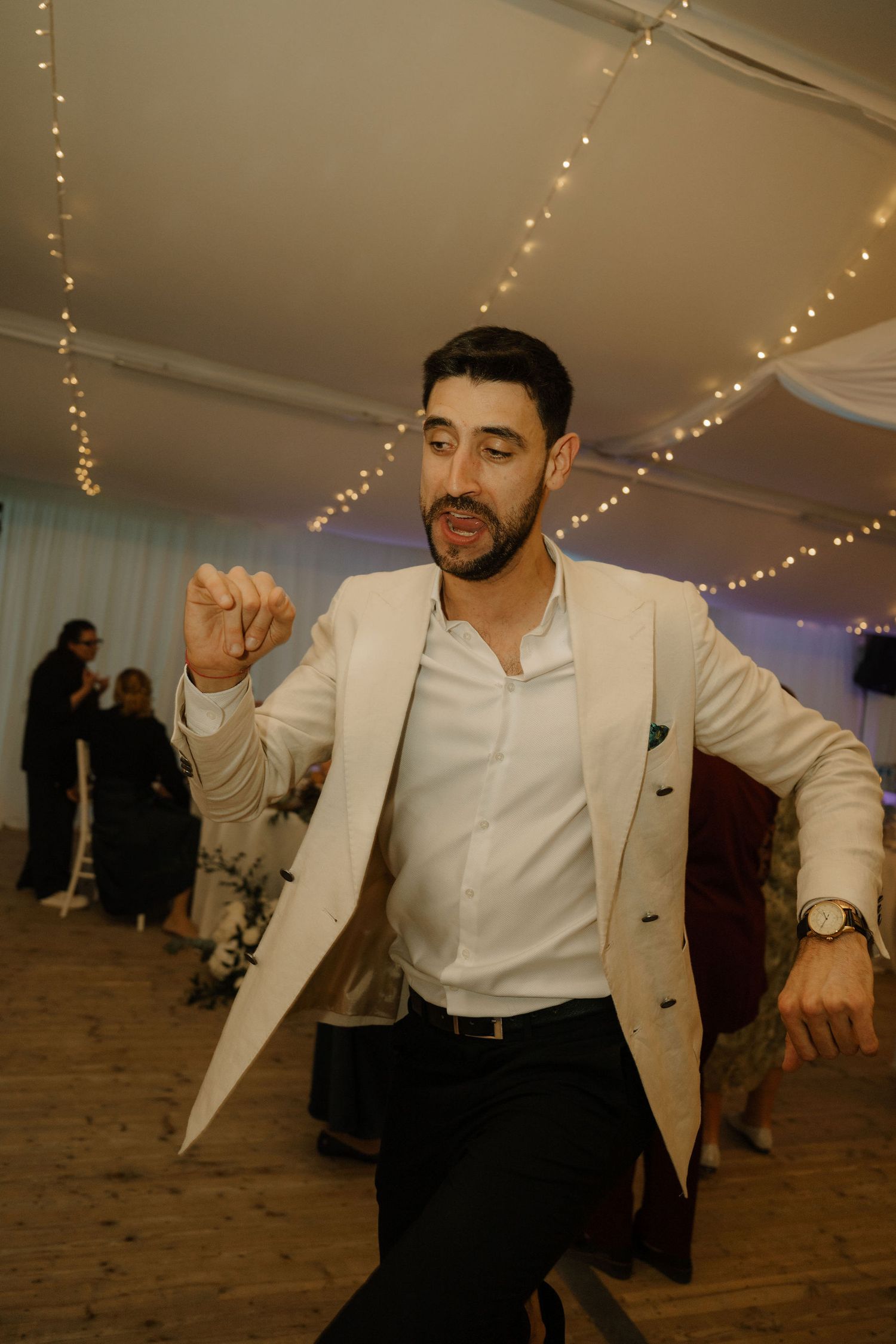 A man in a white blazer and black pants dancing energetically at a wedding reception with fairy lights in the background.
