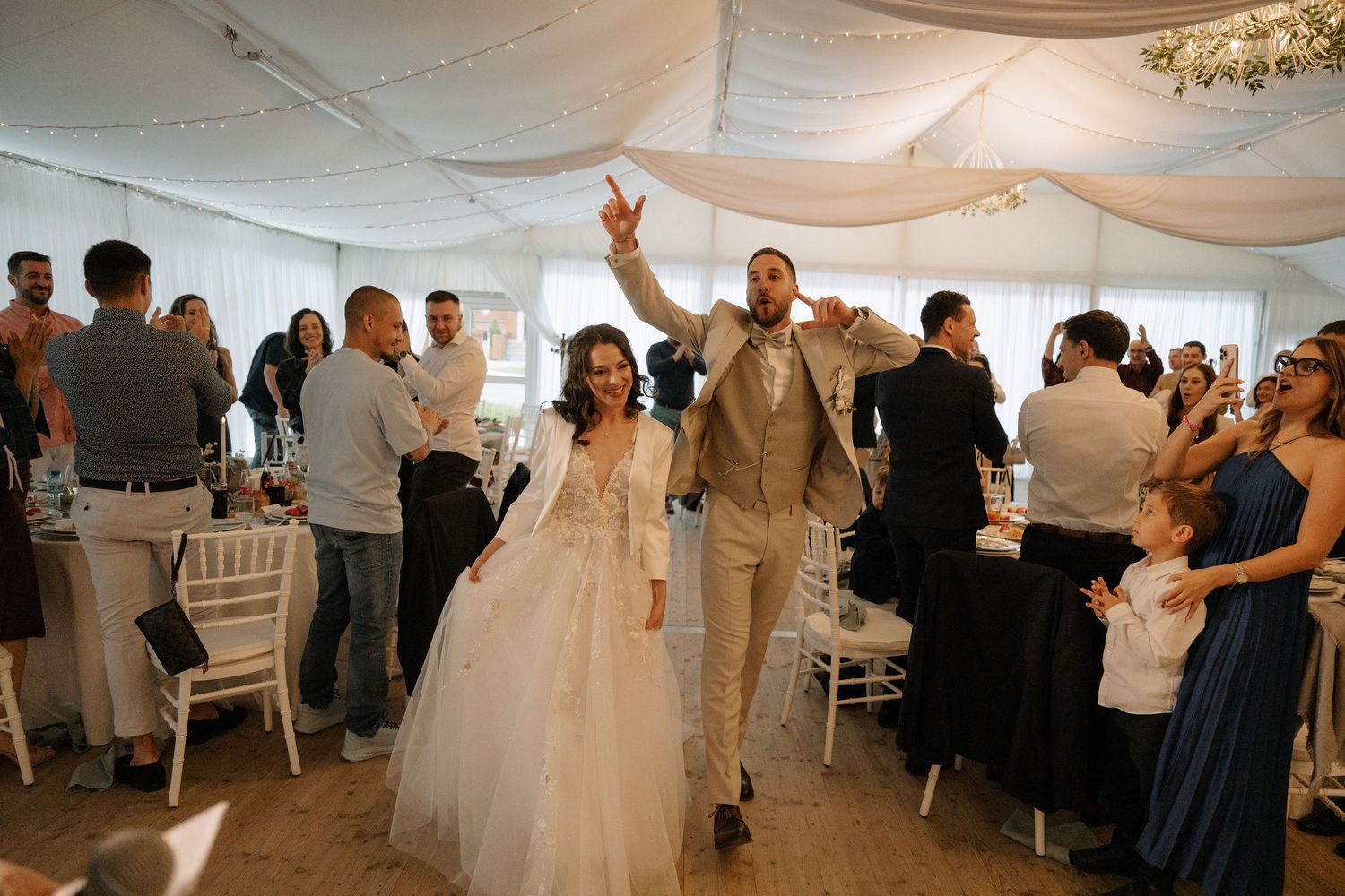 Bride and groom make grand entrance at elegant wedding reception in white draped venue with seated guests.