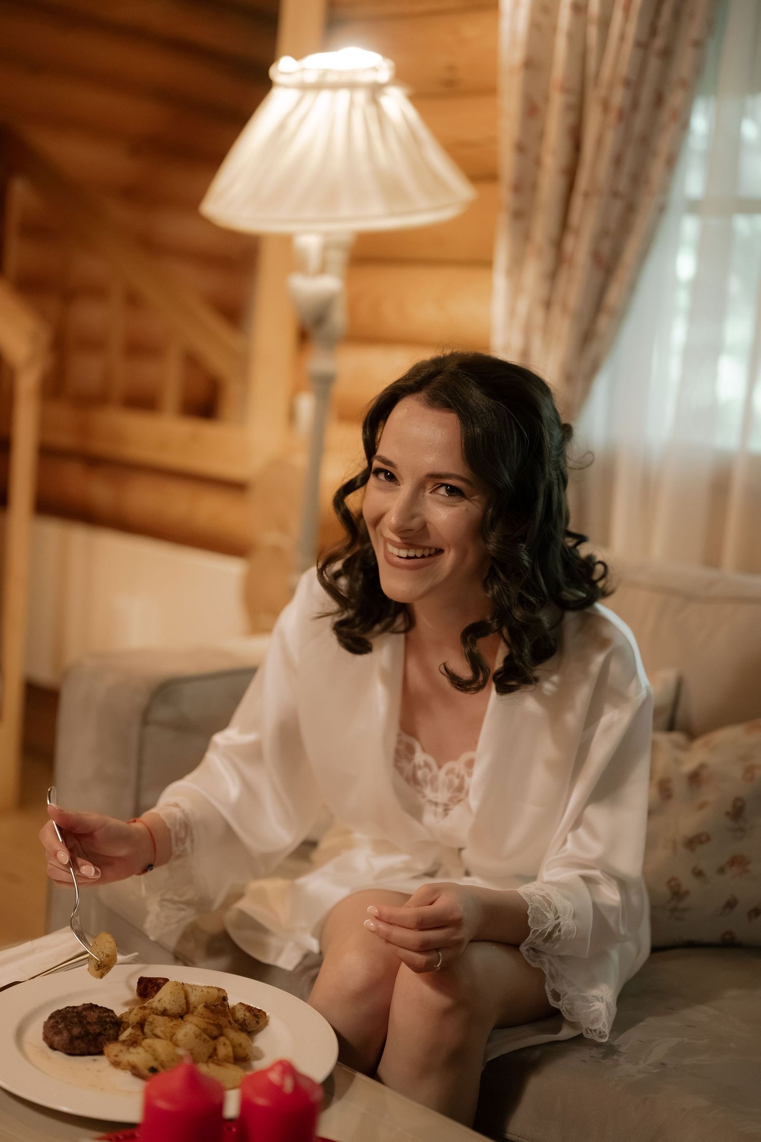 Smiling brunette woman in white robe enjoying breakfast on a cozy sofa in a warm wooden cabin interior.