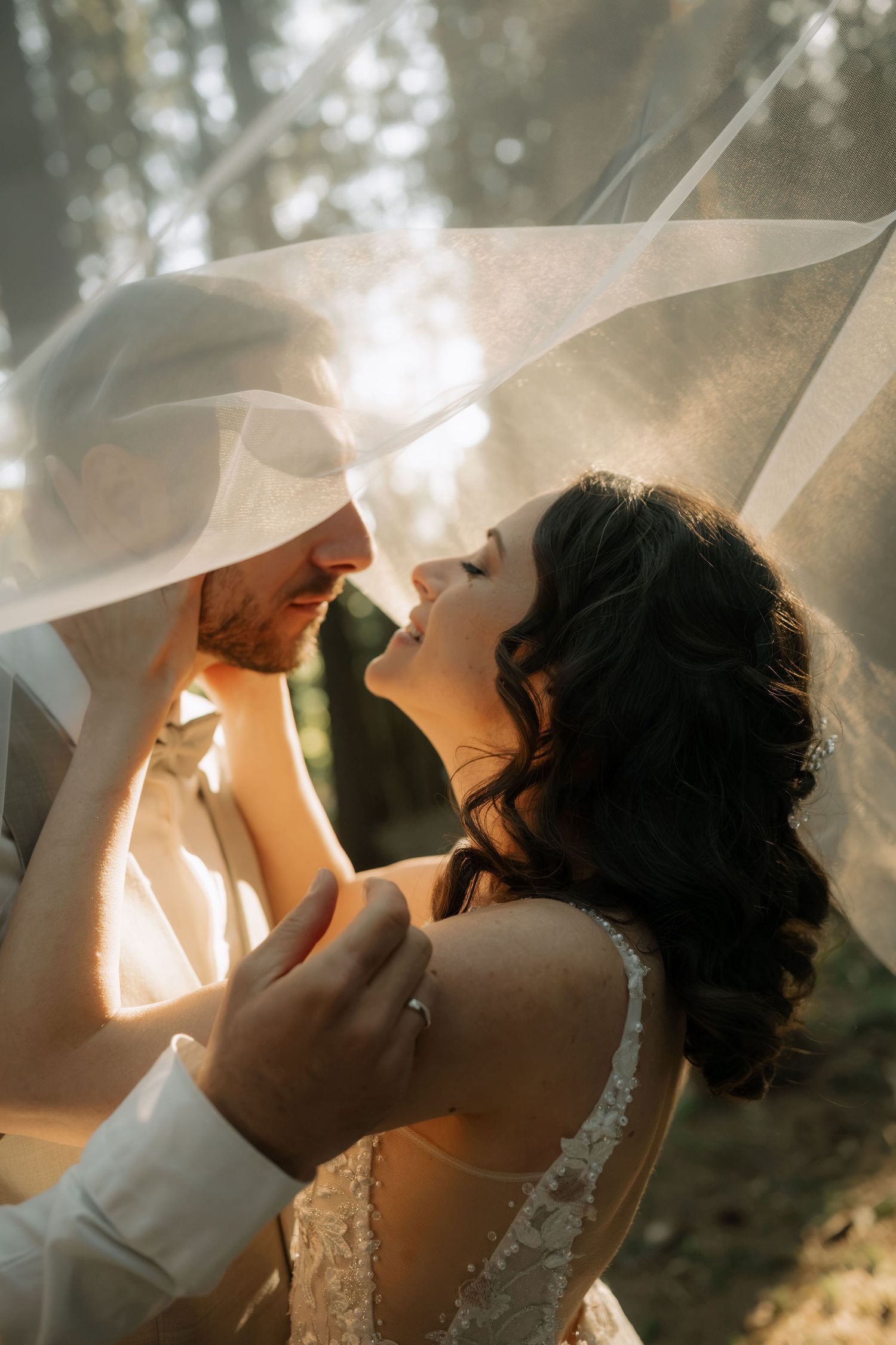 Bride and groom share a romantic moment under a flowing veil, bathed in golden sunlight on their wedding day.