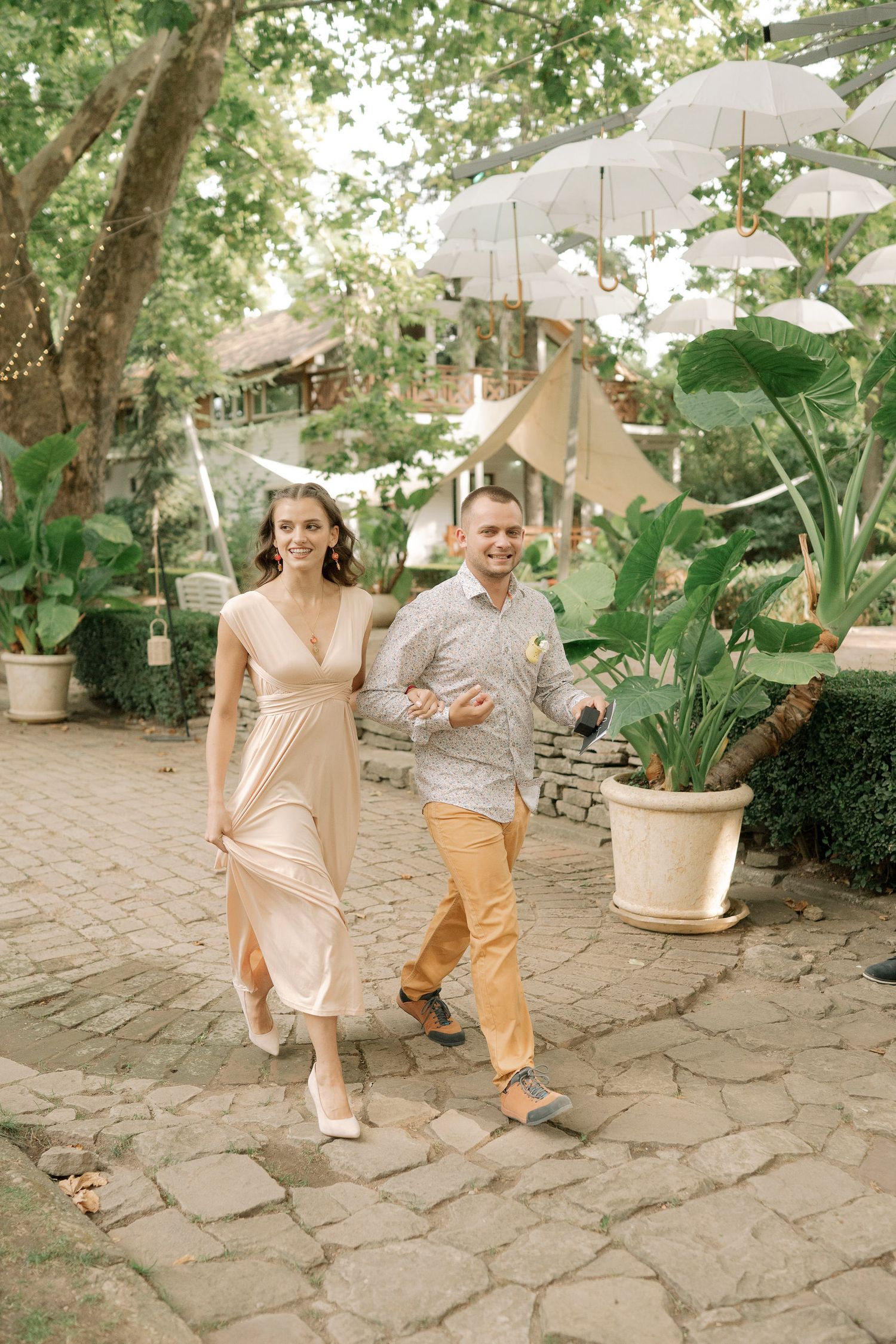 A couple walks together on a stone path under white umbrellas, surrounded by lush greenery and potted plants.