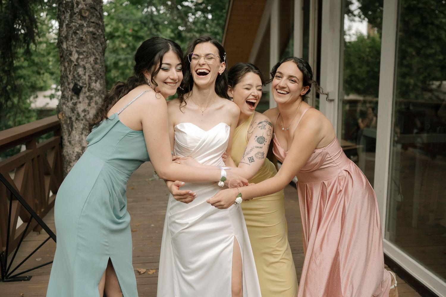 Four women in elegant dresses share a joyful moment of laughter together on a wooden porch.