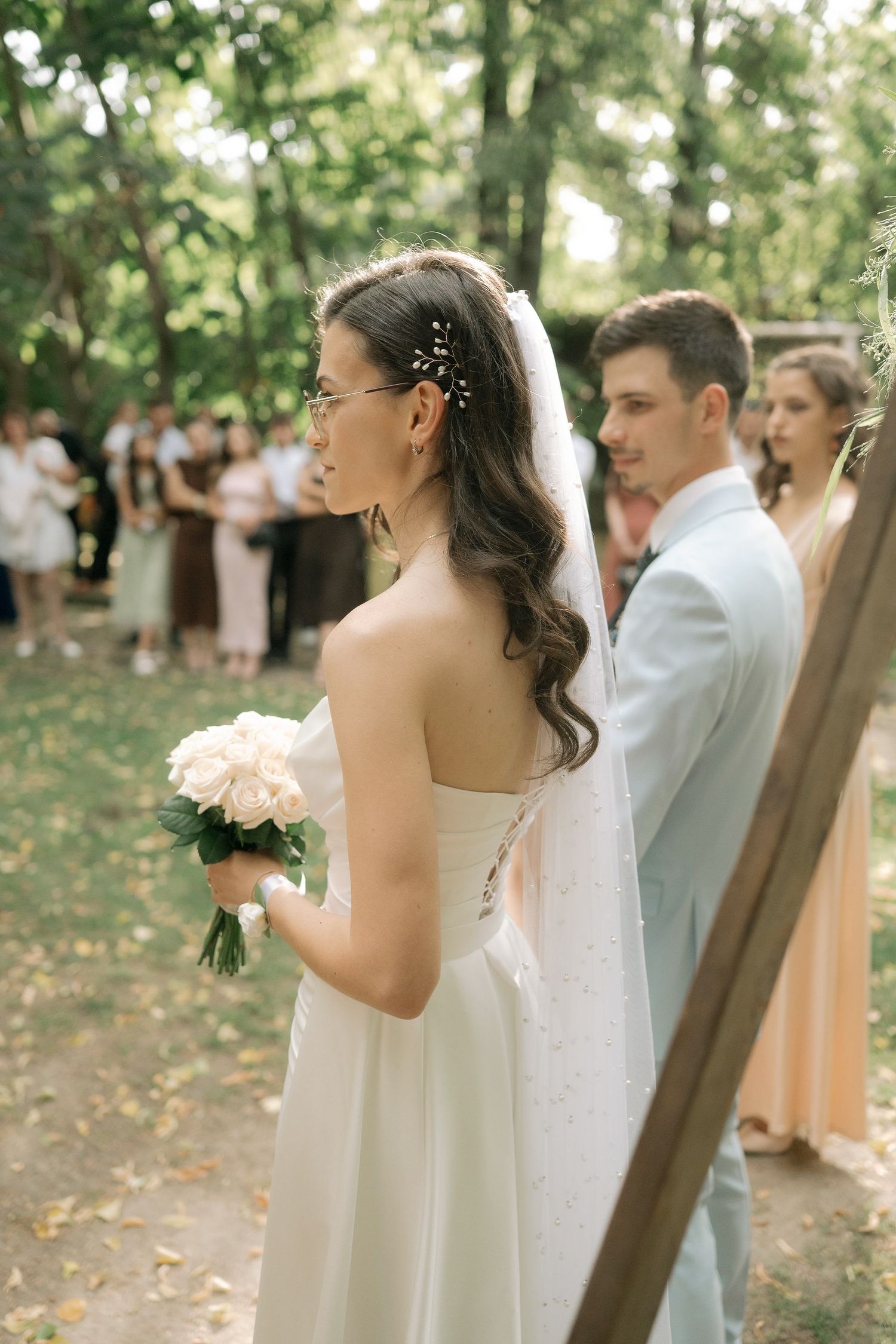 A romantic outdoor wedding ceremony in a wooded setting with guests gathered under trees on a sunny day.