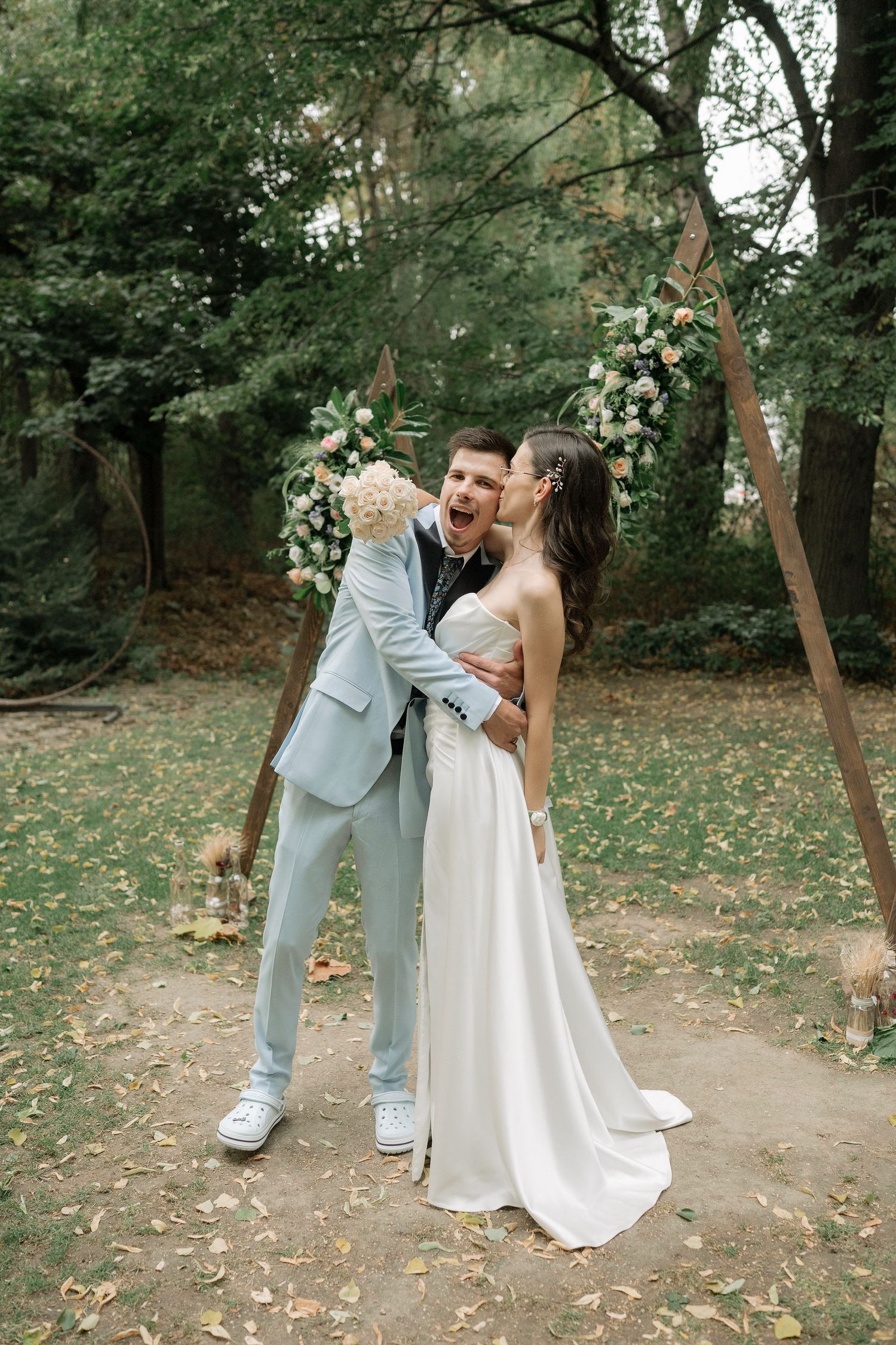 A couple in wedding attire shares romantic moments in a wooded outdoor setting with fallen leaves on the ground.