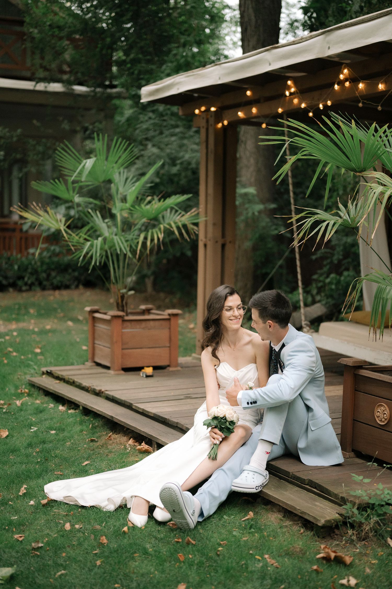 Couple sitting on wooden deck steps surrounded by tropical plants and string lights in garden setting.