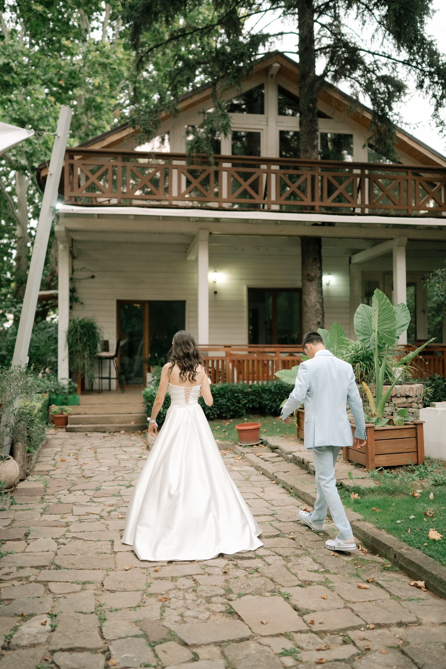 A couple walks along a stone pathway towards a rustic two-story cabin with a wooden balcony surrounded by trees.