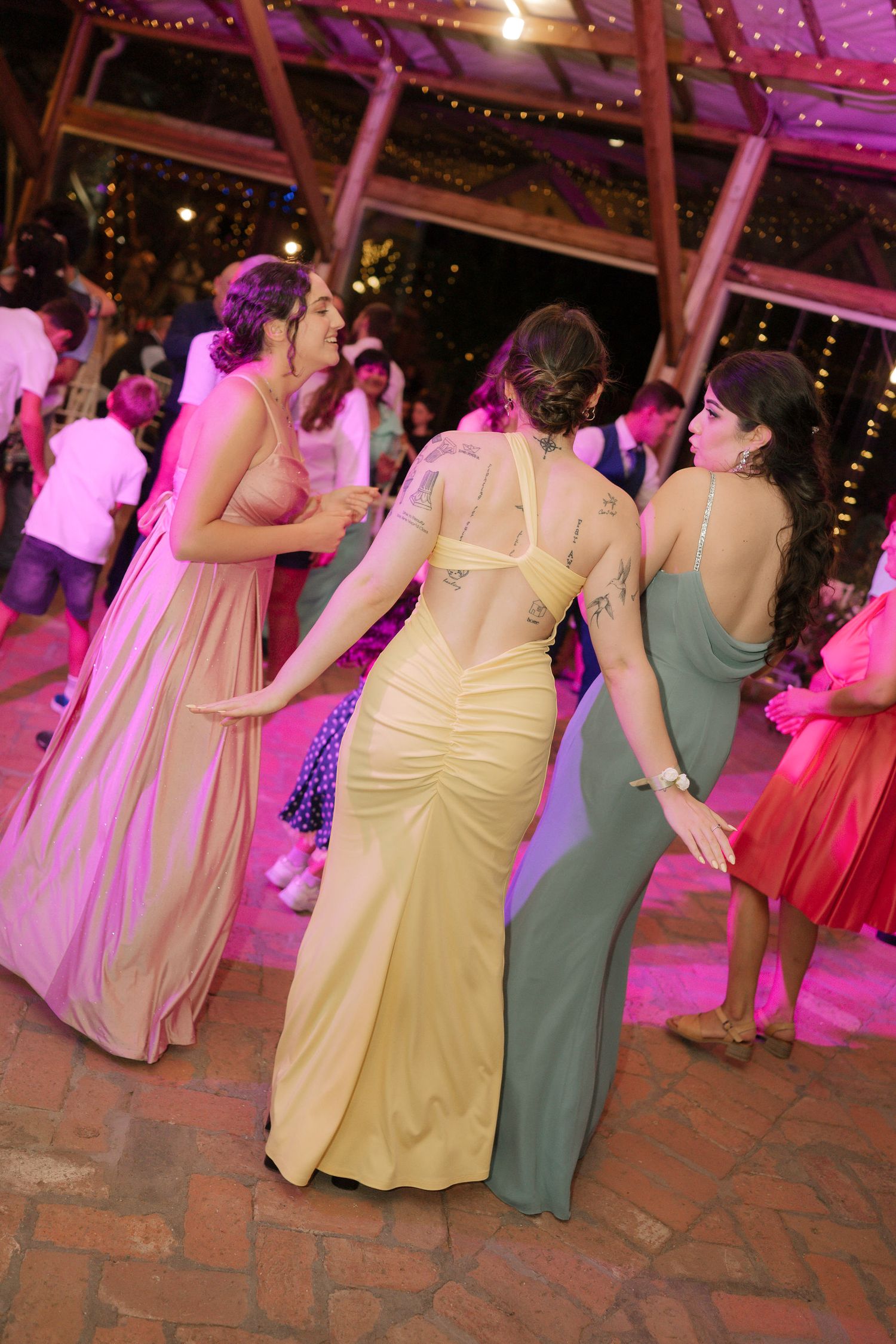 Wedding guests in pastel dresses dance together on a brick patio under pink lighting and twinkling fairy lights.