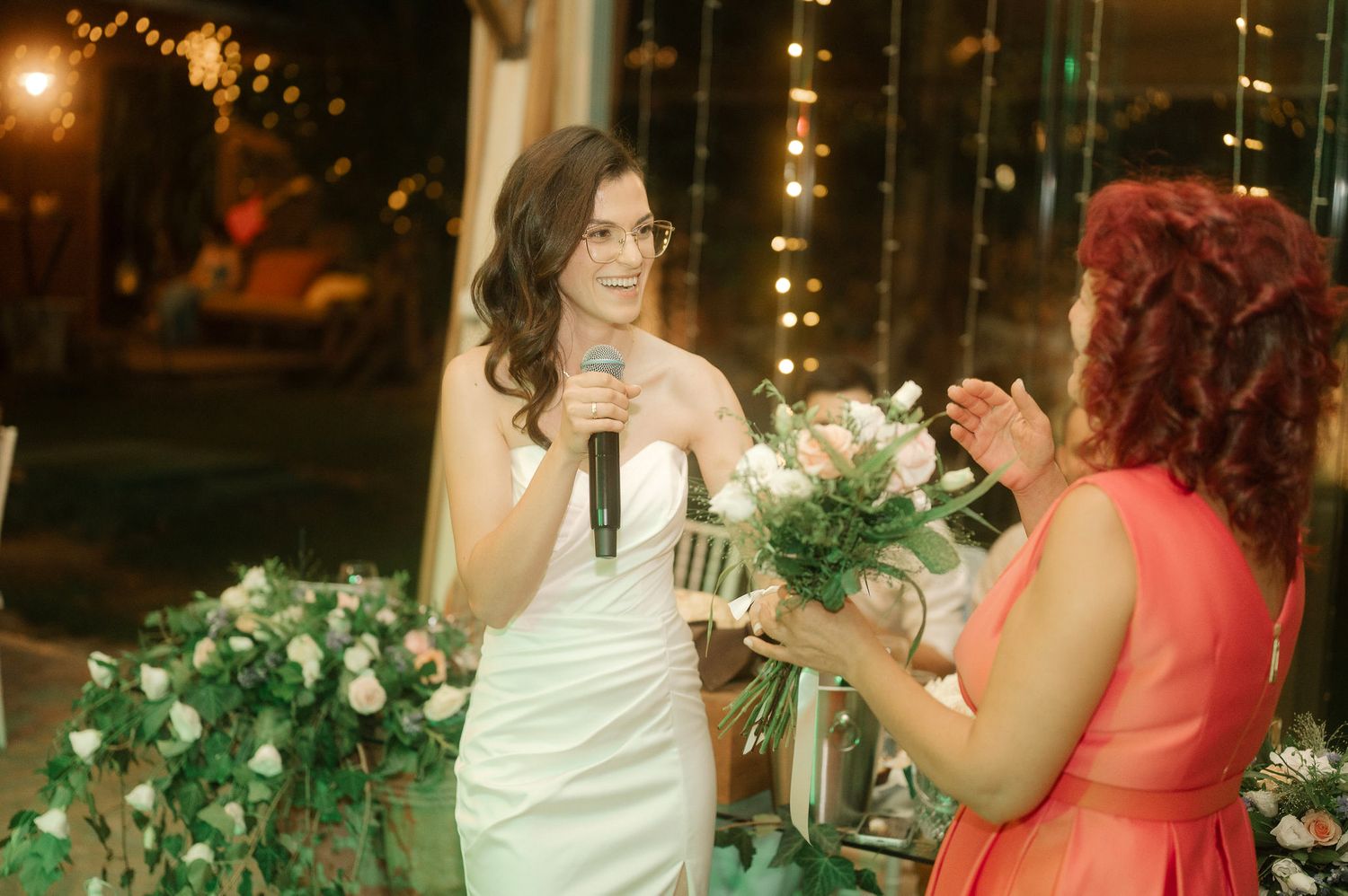 Two people in formal attire share a conversation surrounded by decorative lights and floral arrangements at an evening event.