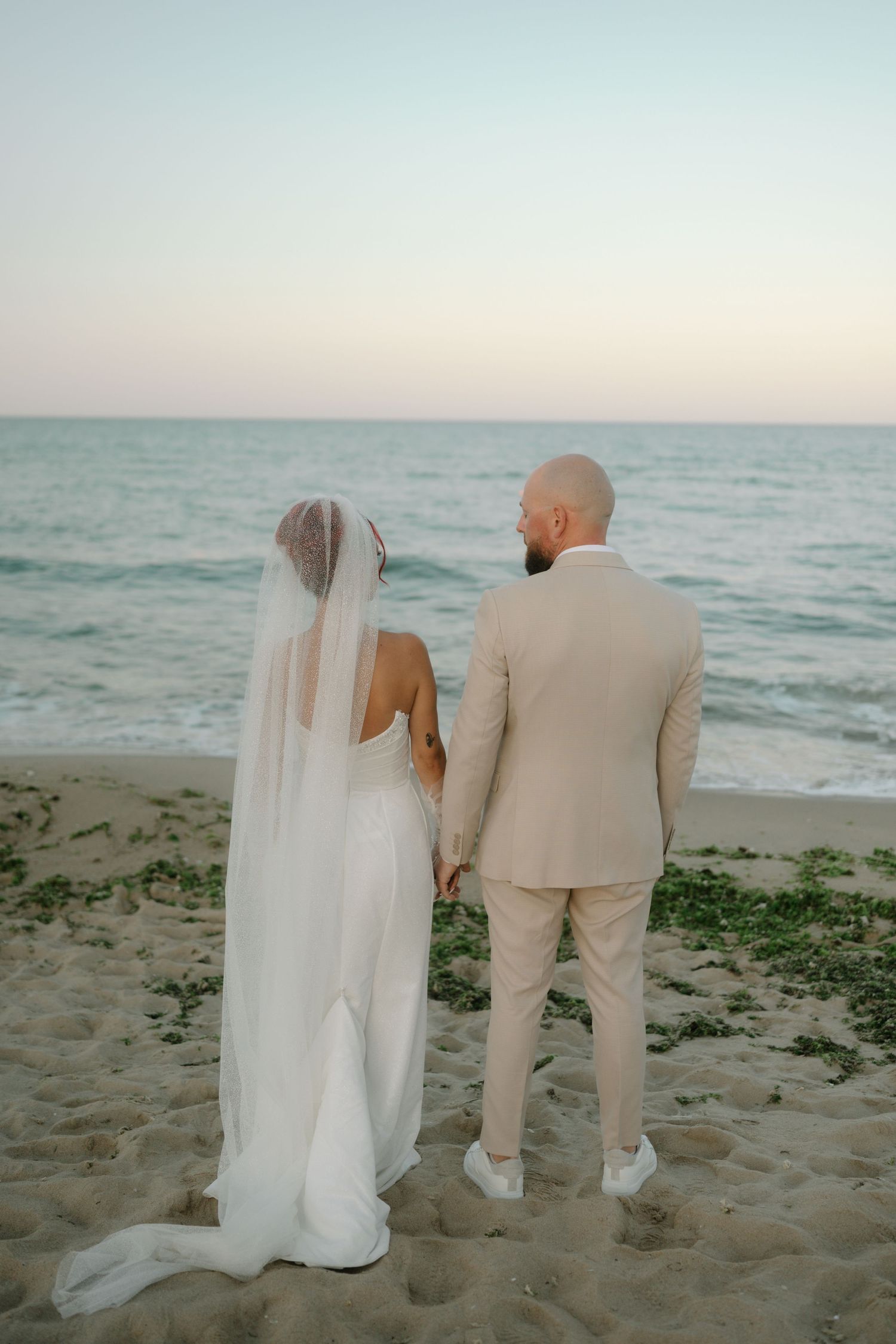 A couple in wedding attire stands together on a sandy beach at sunset looking out at the ocean waves.