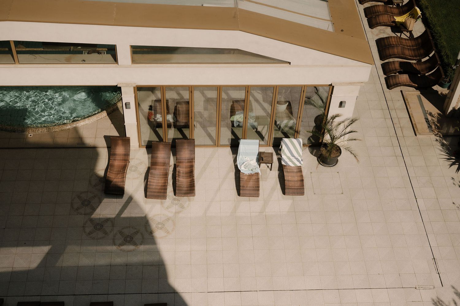 Aerial view of modern wooden lounge chairs arranged along a beige stone patio next to a swimming pool.