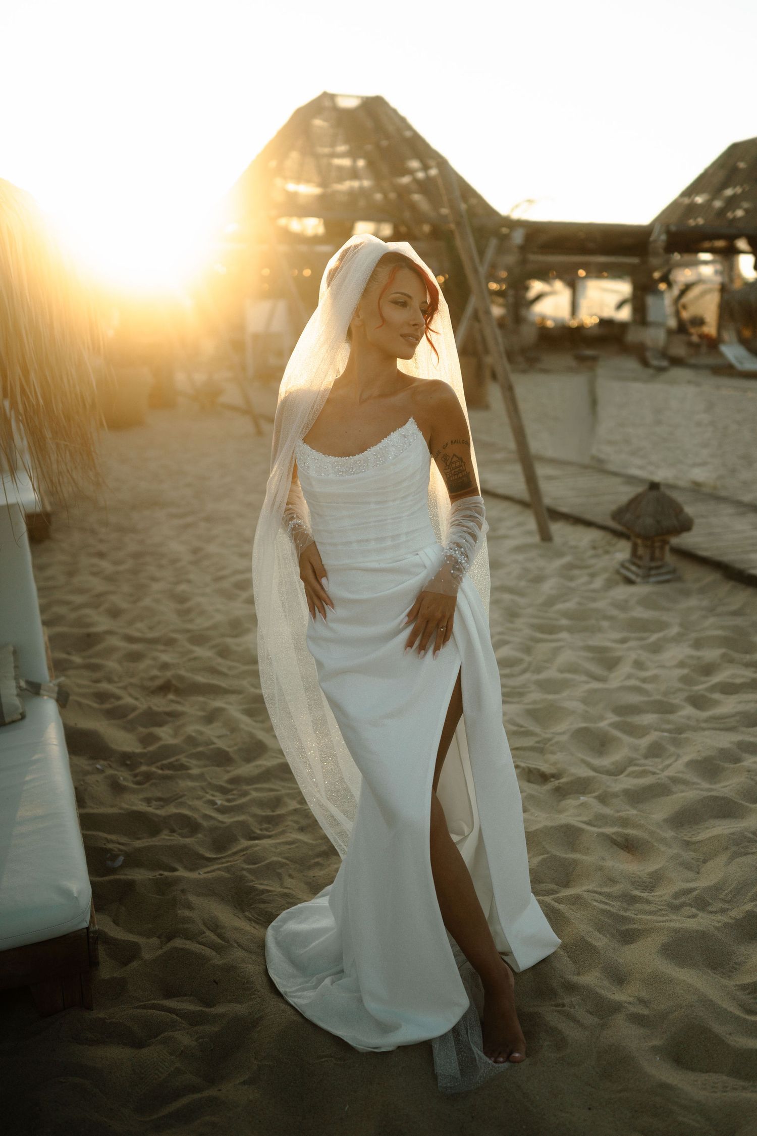 Elegant bride in sleek white wedding dress with flowing veil poses on sandy beach at golden sunset.