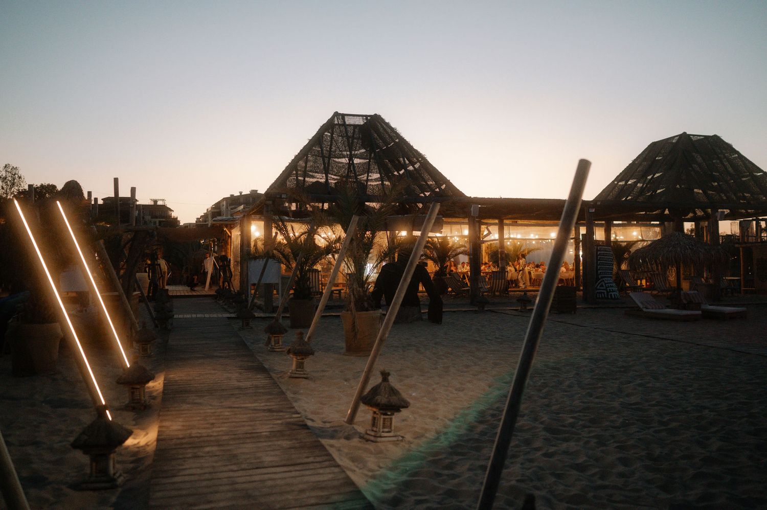 Wooden beach pier extends into the ocean at sunset with silhouetted structures and palm trees against orange sky.