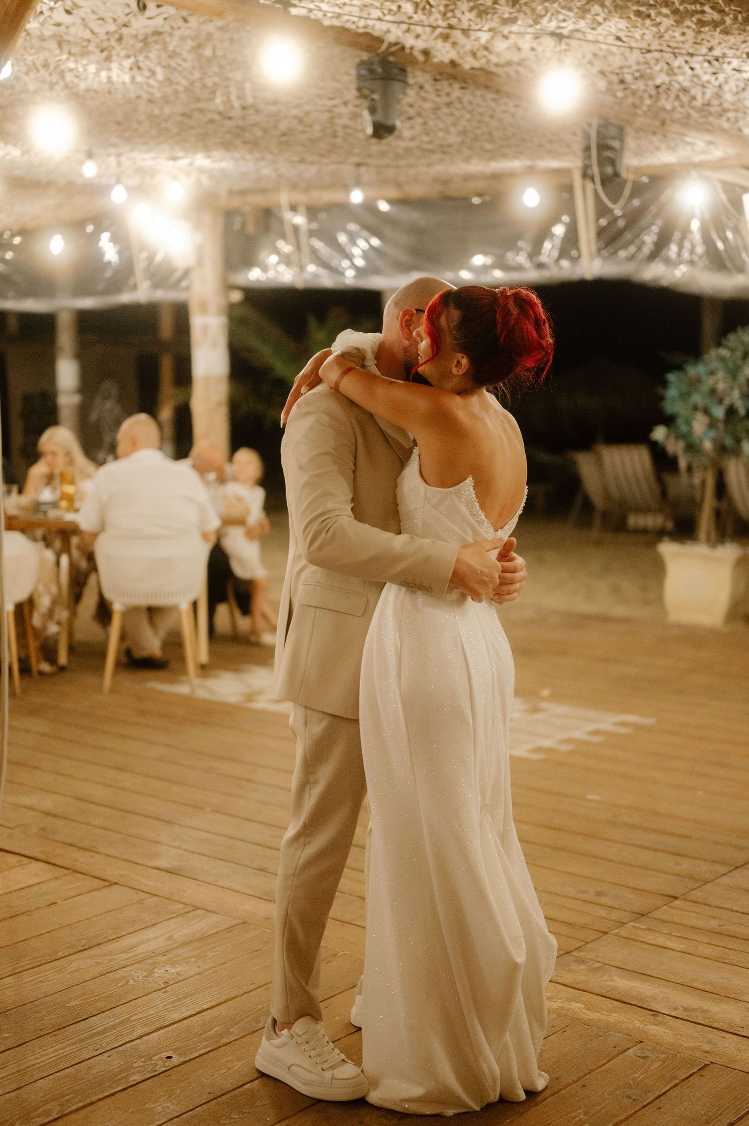 A romantic slow dance under twinkling lights at an outdoor wedding reception on a wooden deck.