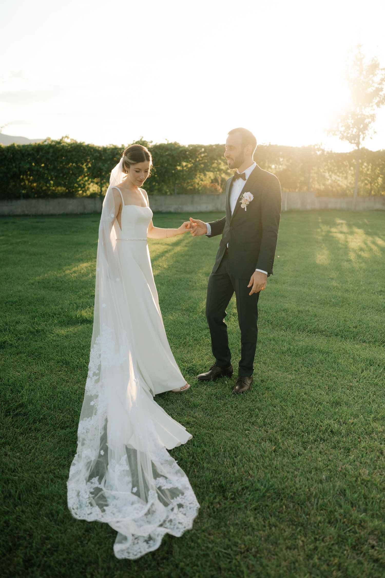 A wedding couple stands on a sunlit lawn with the bride's lace train flowing behind her elegant white gown.