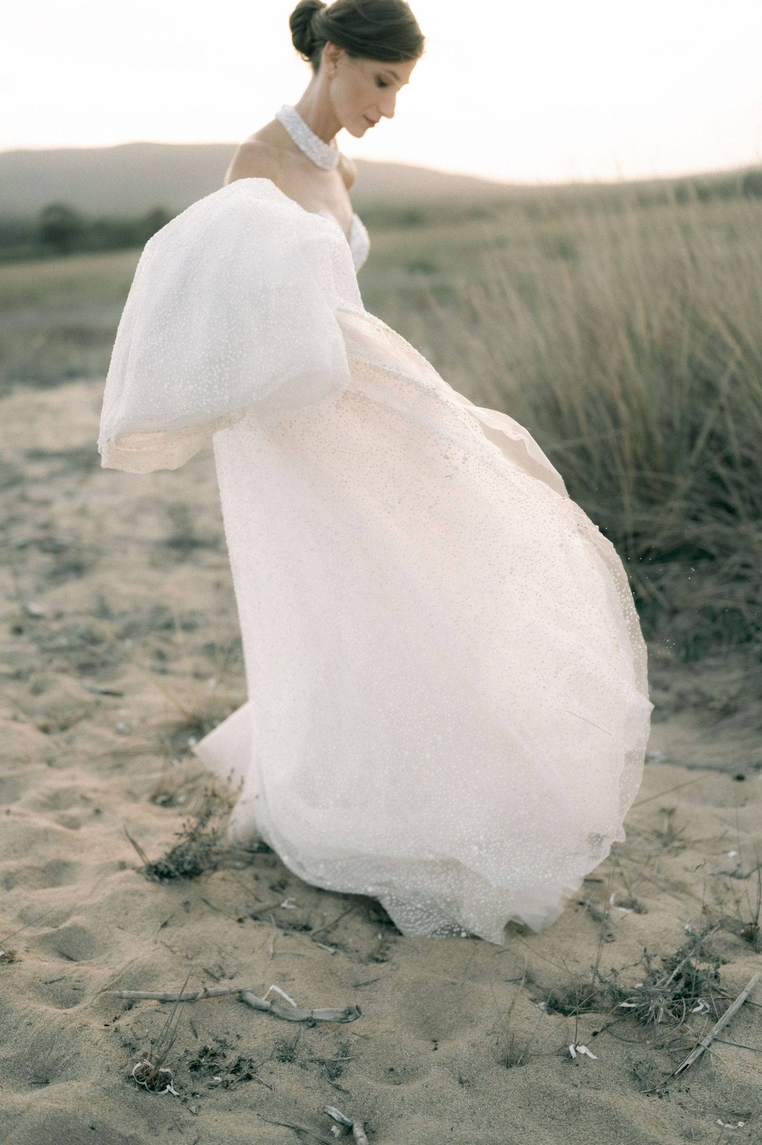 Bride in flowing white lace wedding gown with puff sleeves standing on sandy beach at dusk.