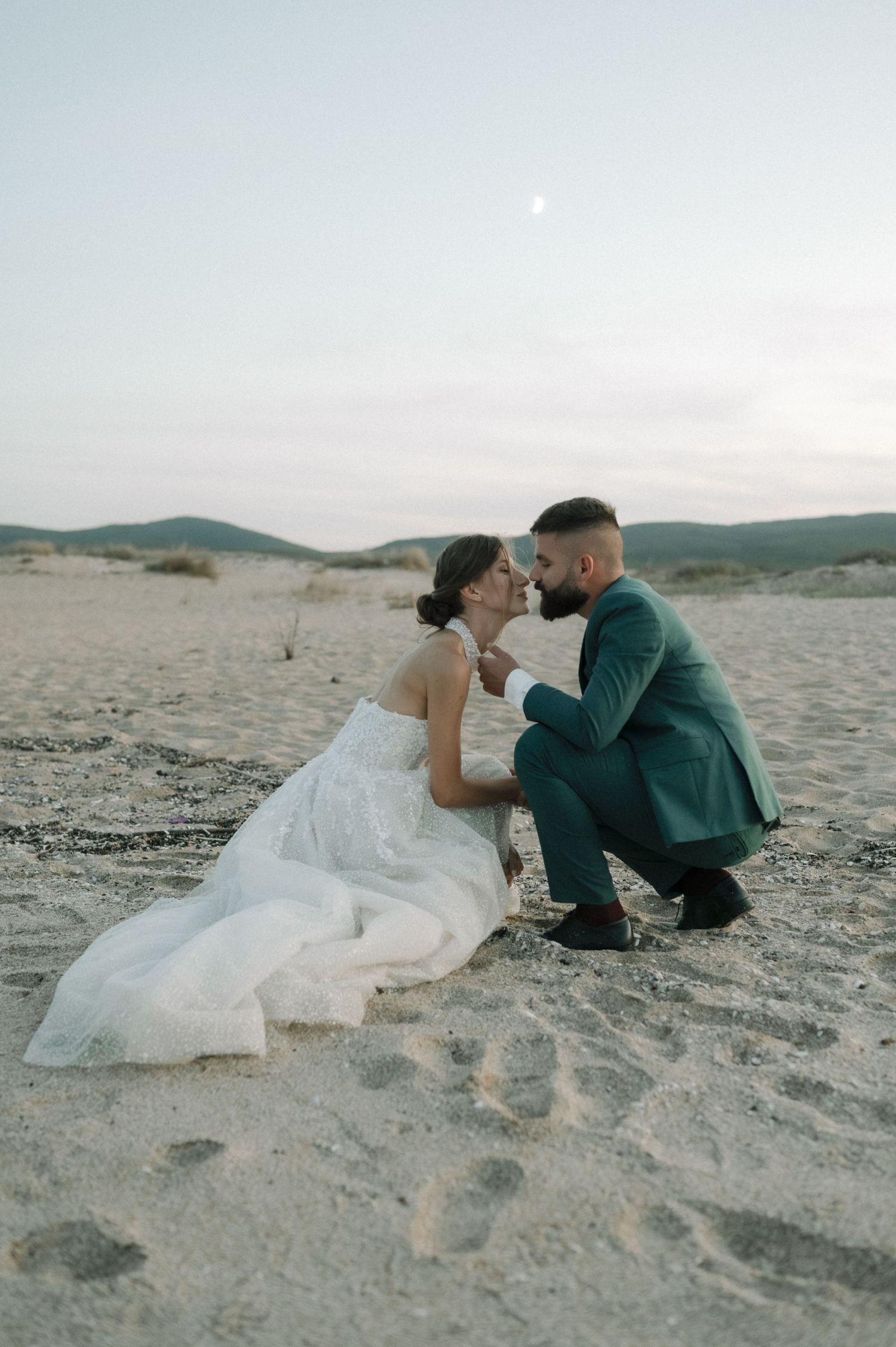 A bride and groom share a romantic moment on a sandy beach at dusk, dressed in a white gown and teal suit.