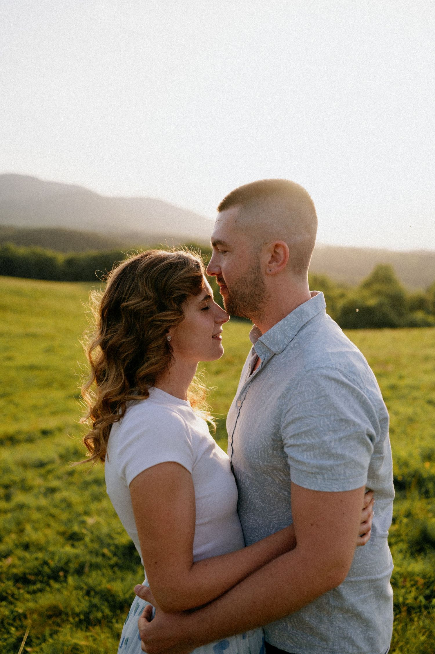 A romantic couple embraces while standing in a sunlit field with mountains visible in the distance.