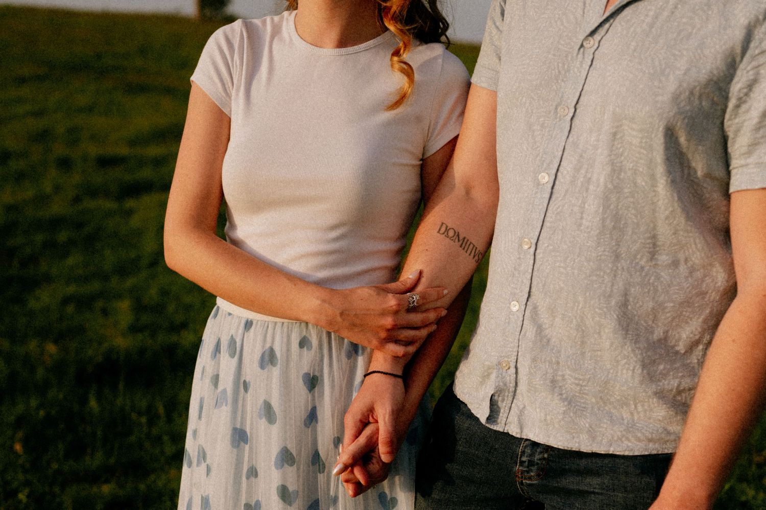 A couple stands together in a grassy field during golden hour with power lines visible in the background.