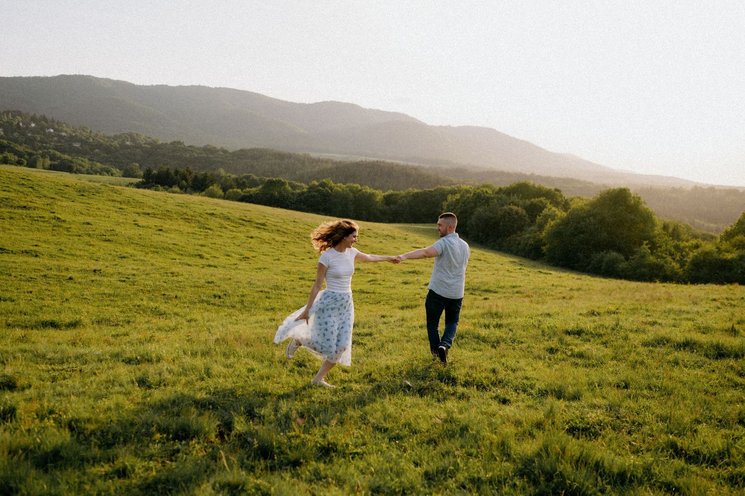 Couple strolls together across a verdant hillside with mountains in the distance.