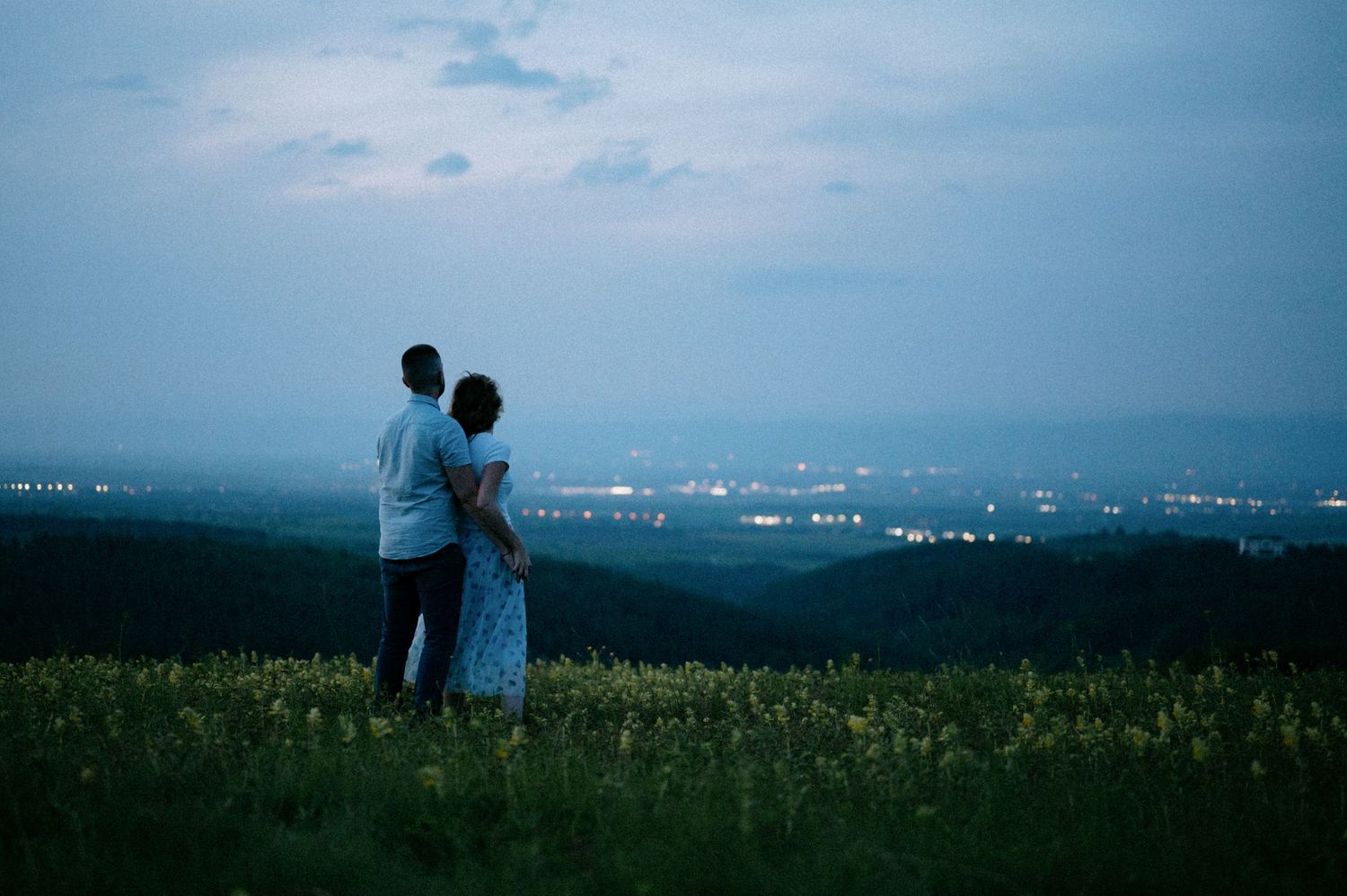 A couple stands together on a scenic overlook with city lights twinkling in the distance.