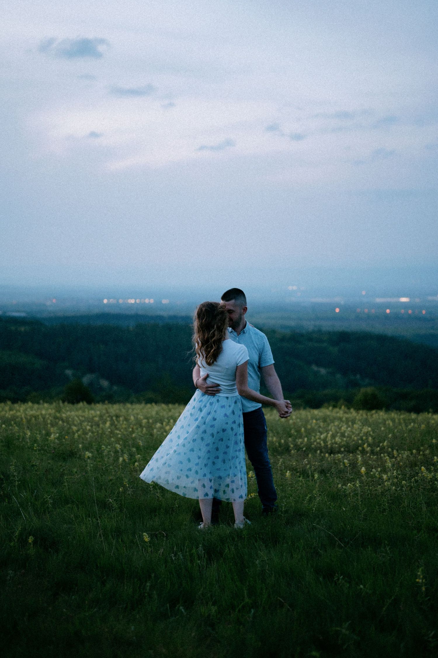 A couple shares a romantic moment in a meadow with city lights visible in the distance.