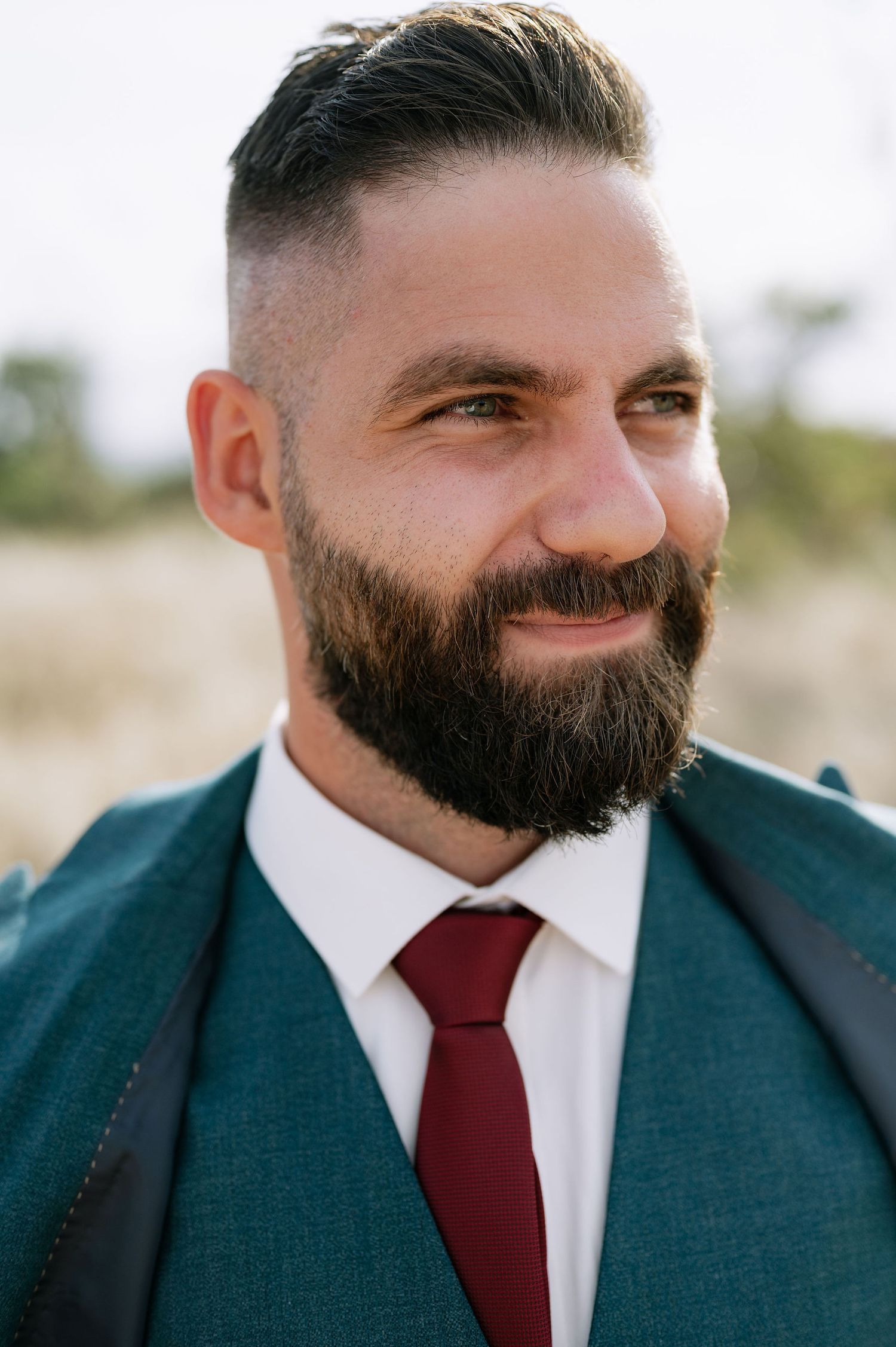 A close-up portrait showing a dark beard and burgundy tie against a navy suit.