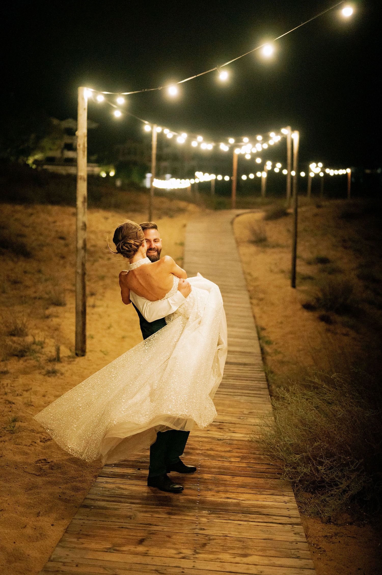 Newlyweds share an intimate moment on a wooden boardwalk lit by overhead string lights.