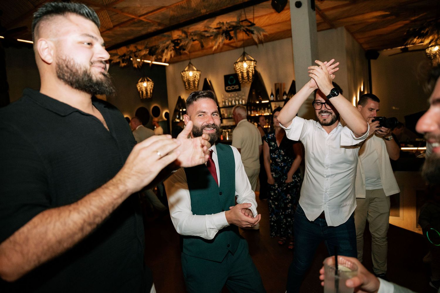 Wedding guests dancing and celebrating with drinks at an indoor reception with rustic wooden ceiling and pendant lighting.