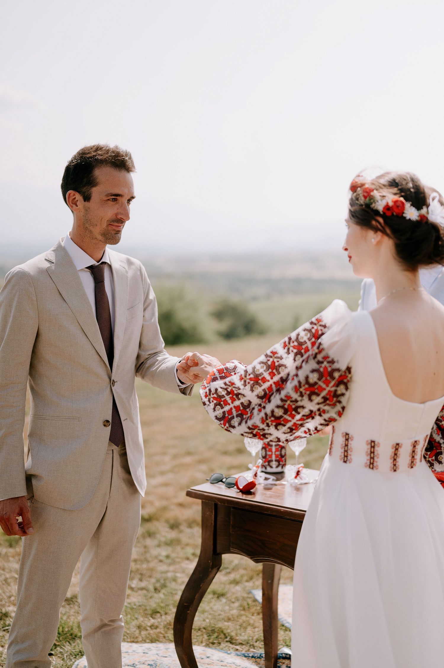A couple in wedding attire stands in a desert landscape with a wooden table and red floral arrangements.