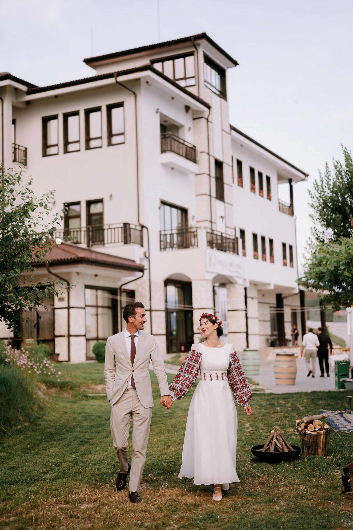 A couple in wedding attire walking on a grassy hillside by a white building, wearing a beige suit and white dress.