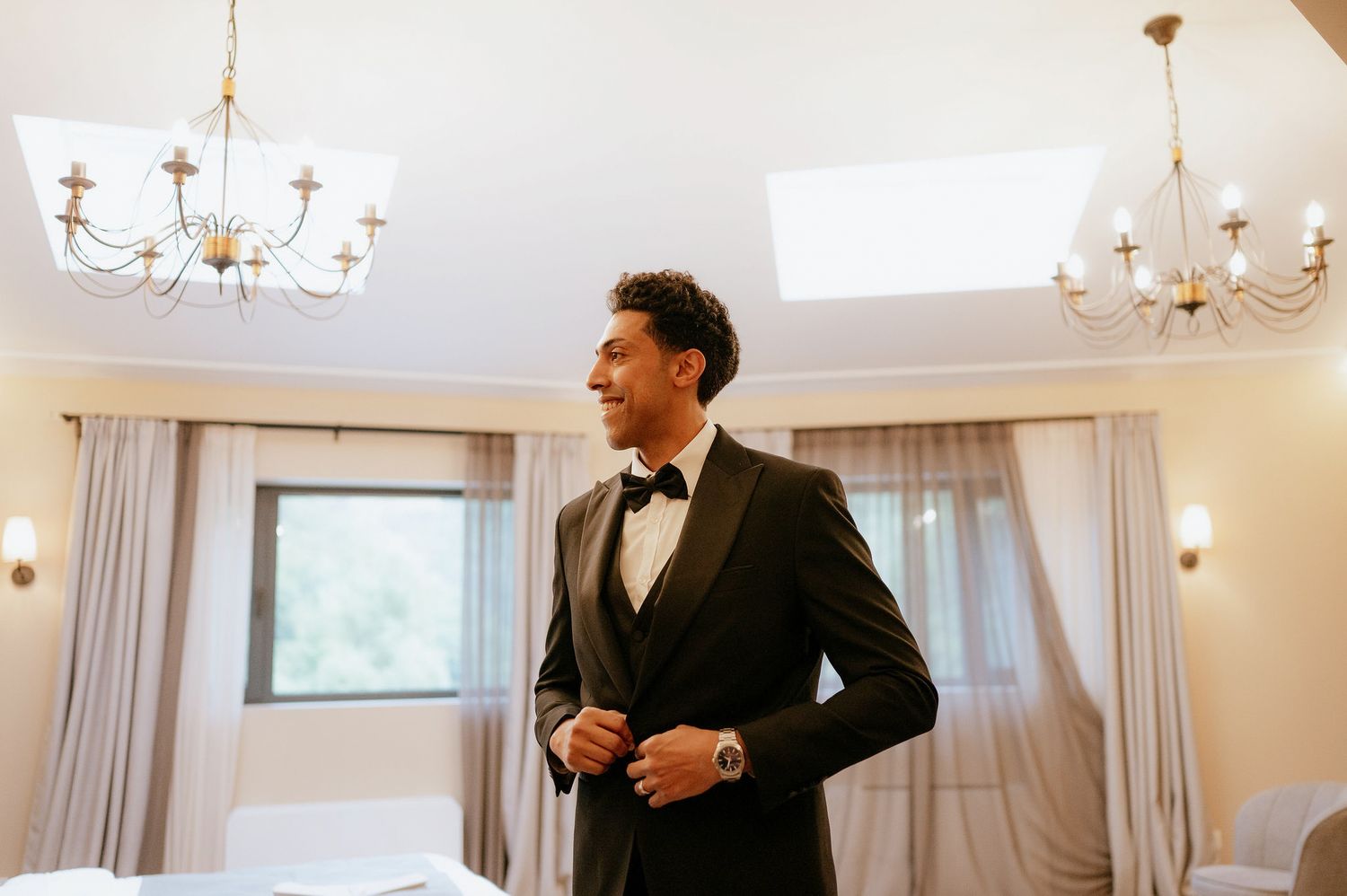 Wedding preparation scene in bright room with chandelier and flowing curtains.