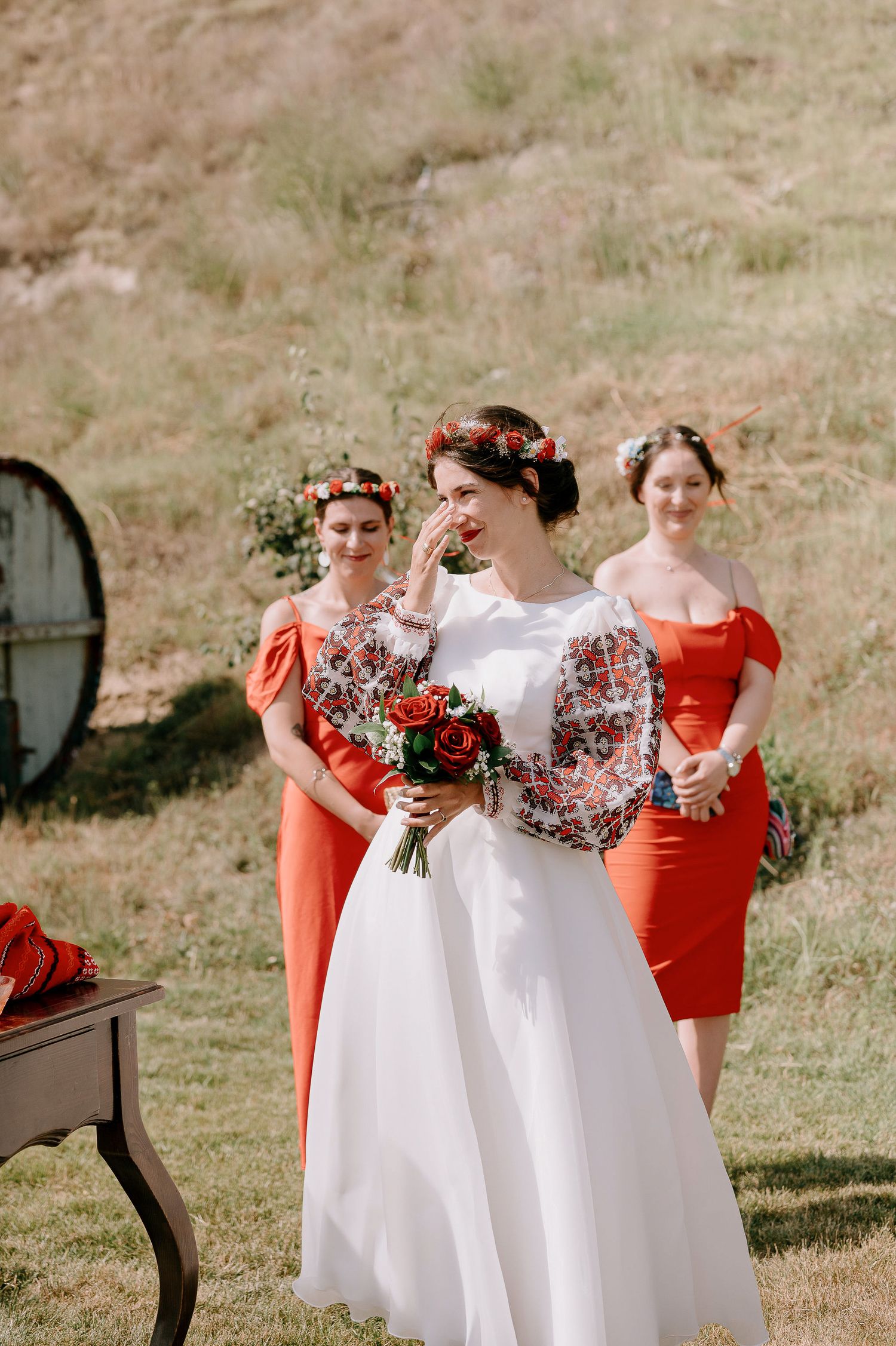 Bridesmaids in coral red dresses pose with bride in white gown against natural landscape backdrop.