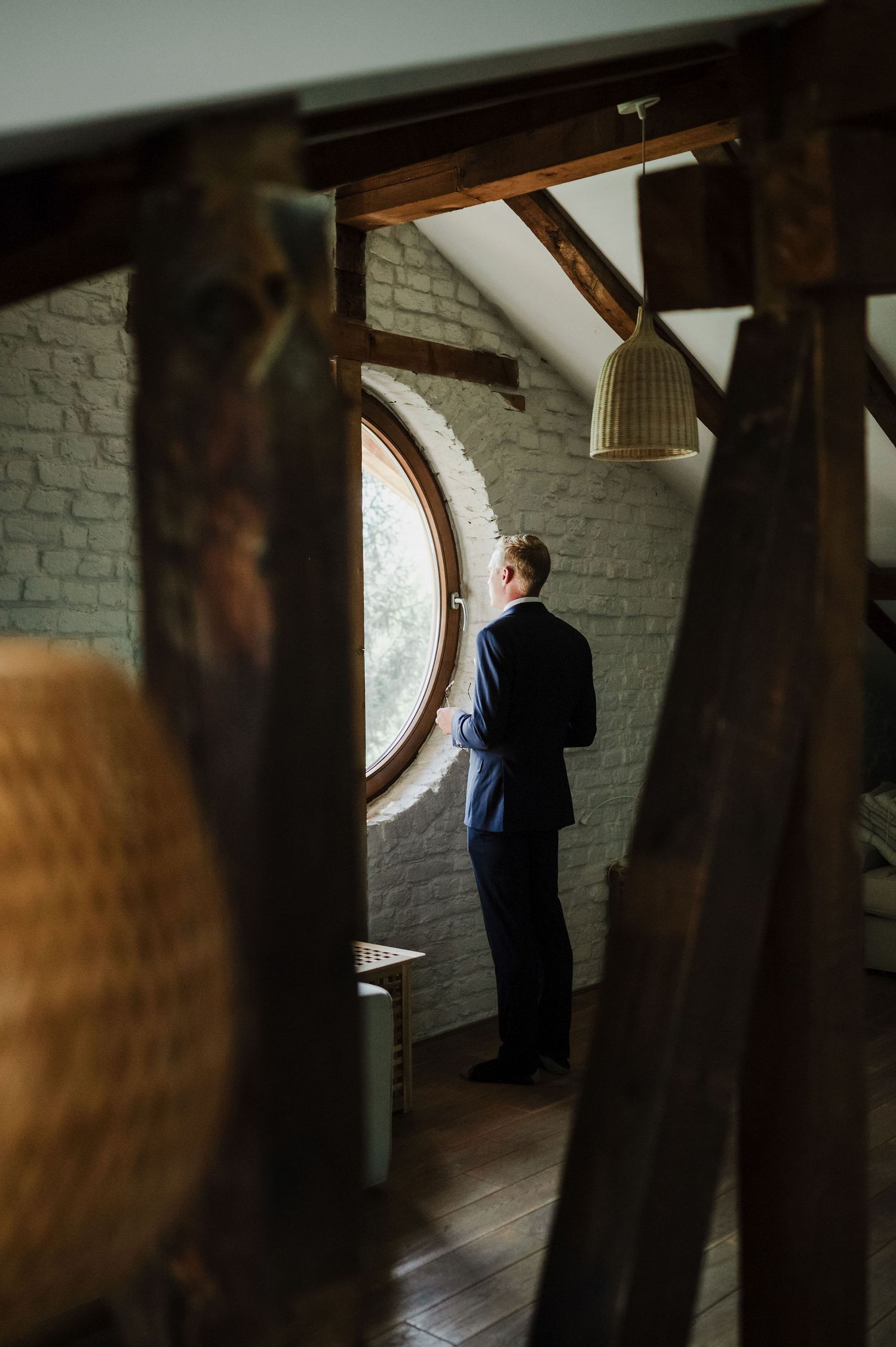 An atmospheric view of a person standing by a round window in a rustic attic space.