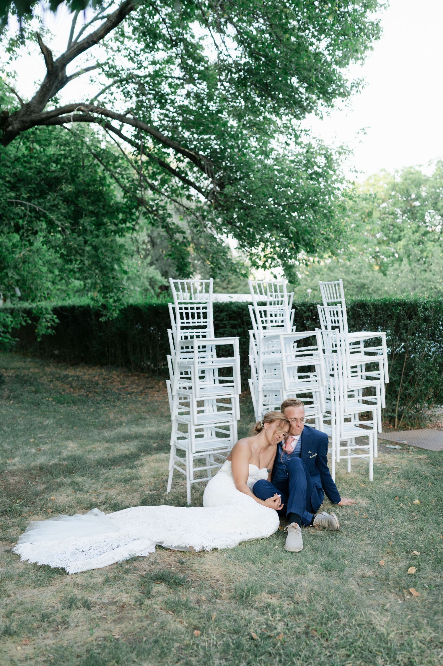 A romantic wedding couple poses on grass near white Chiavari chairs in an outdoor garden setting.