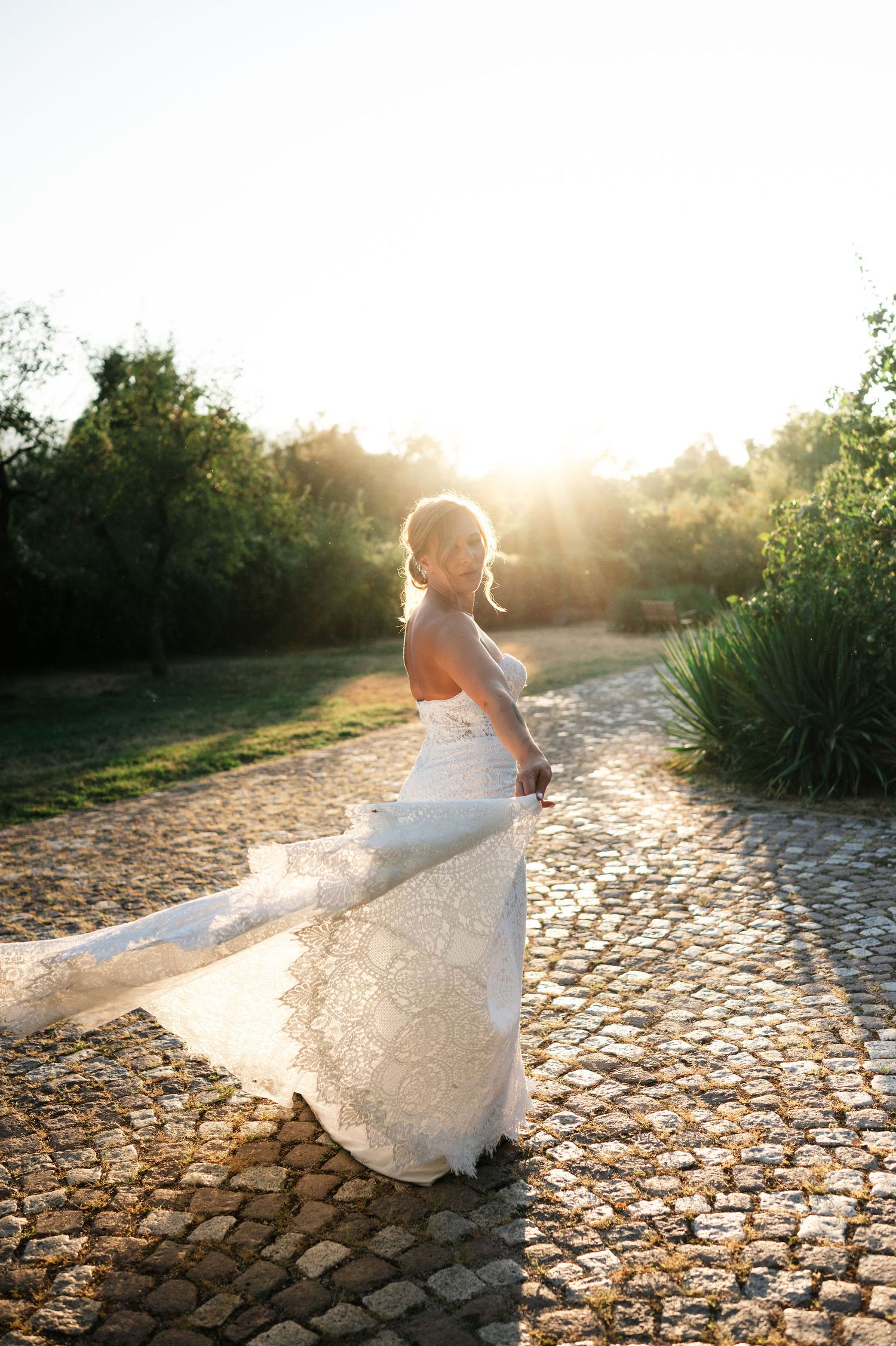A white wedding dress flows gracefully on a cobblestone path during golden hour sunset.