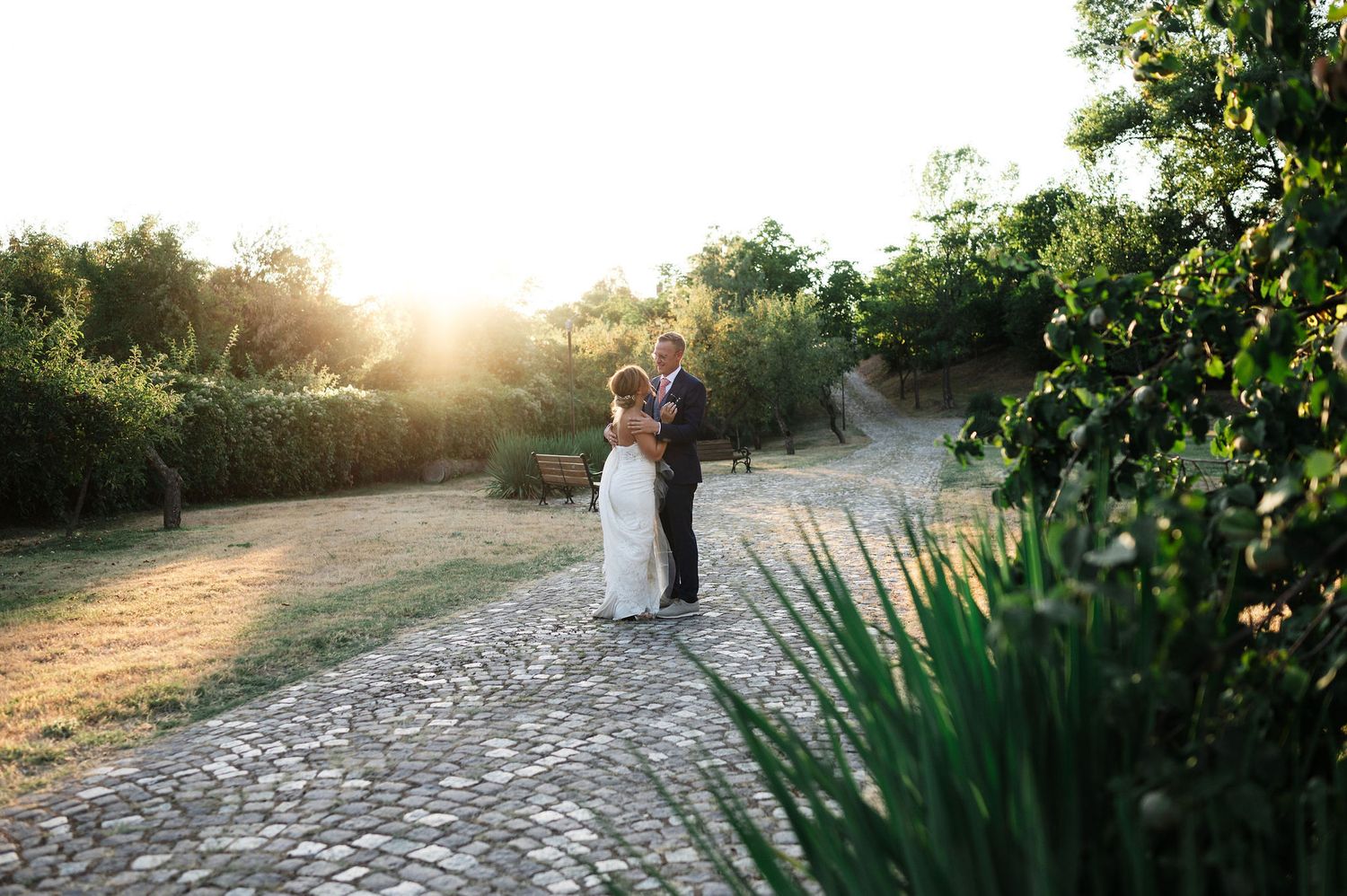 Romantic wedding photo of a couple embracing on a stone pathway surrounded by greenery at sunset.