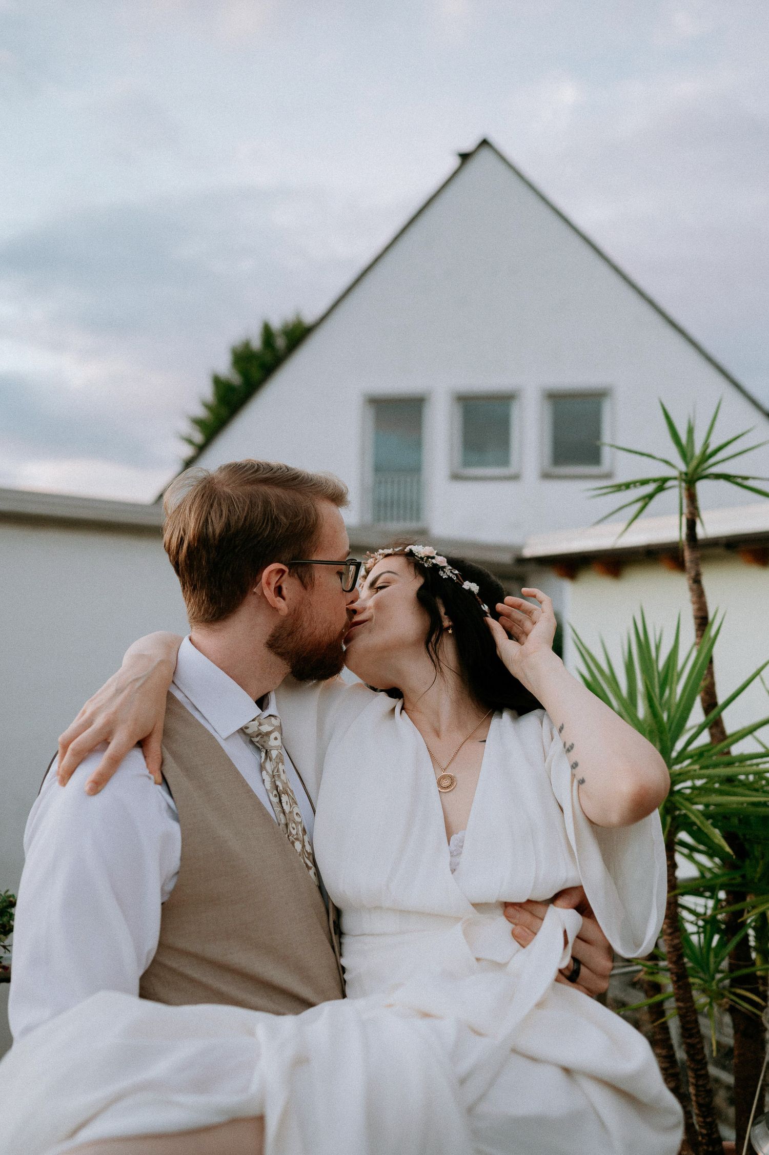 A romantic kiss between newlyweds in front of a white house with tropical plants.