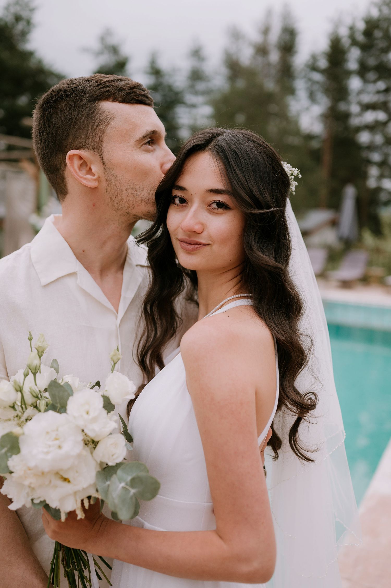 Intimate wedding portraits captured poolside with white floral bouquet and evergreen backdrop.