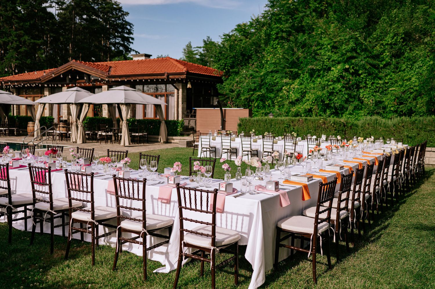 Outdoor wedding dining setup with long tables and dark wooden chairs against a lush green backdrop.