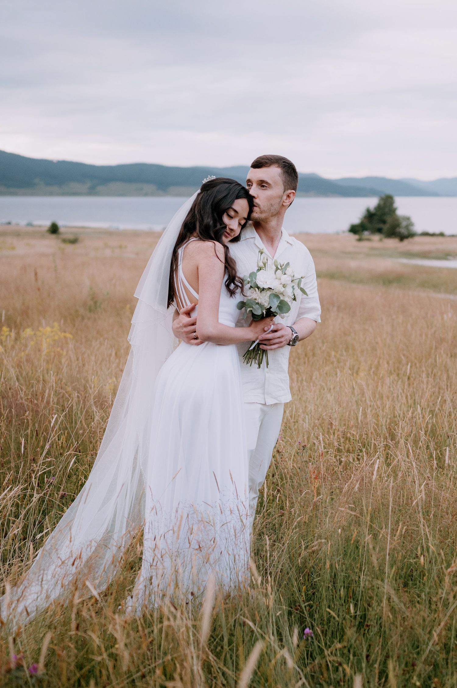 Wedding couple shares tender moments in a serene meadow with mountains and lake views.