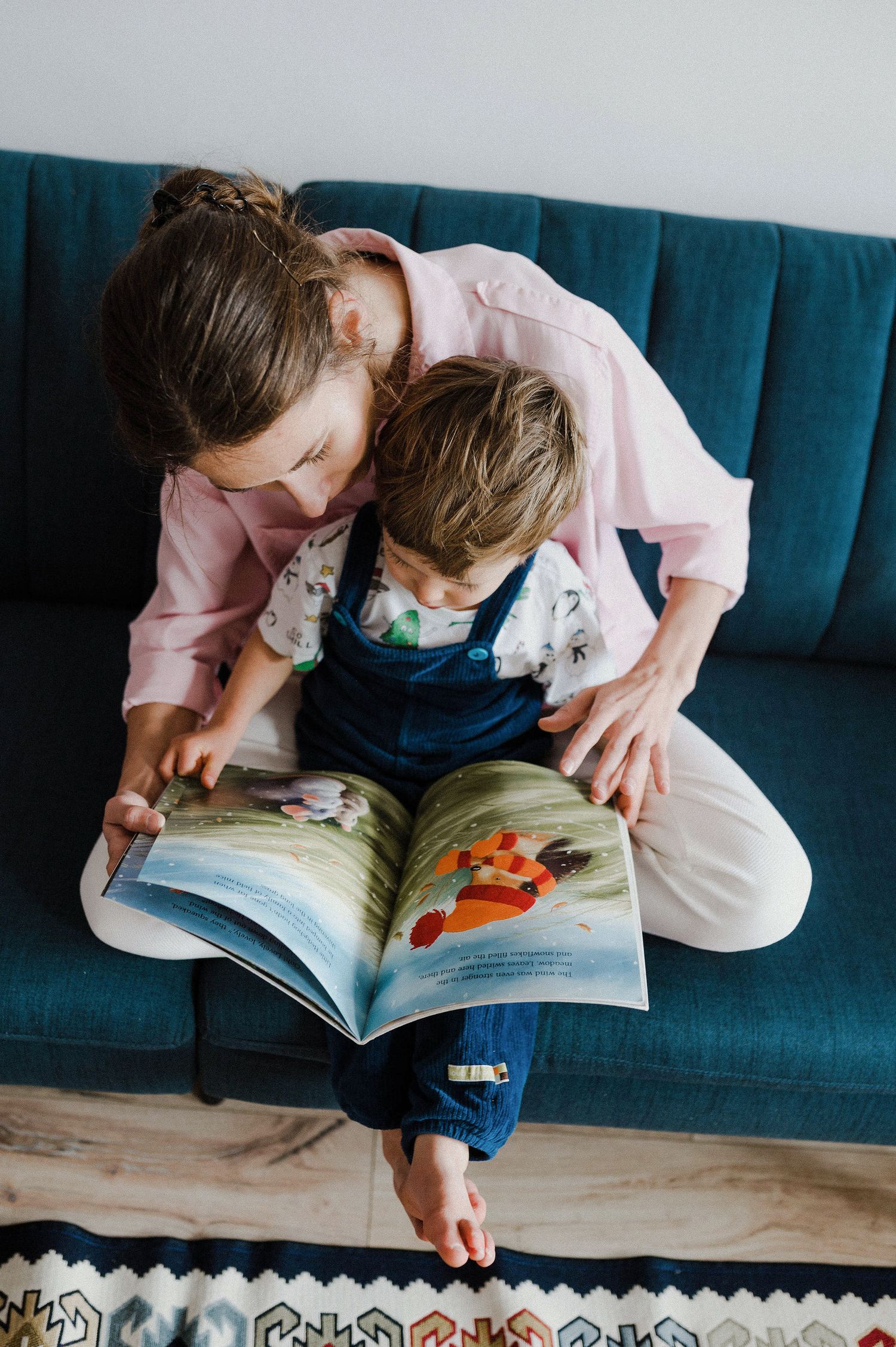 A child in pink sits on a blue couch reading a picture book with intense focus and engagement.