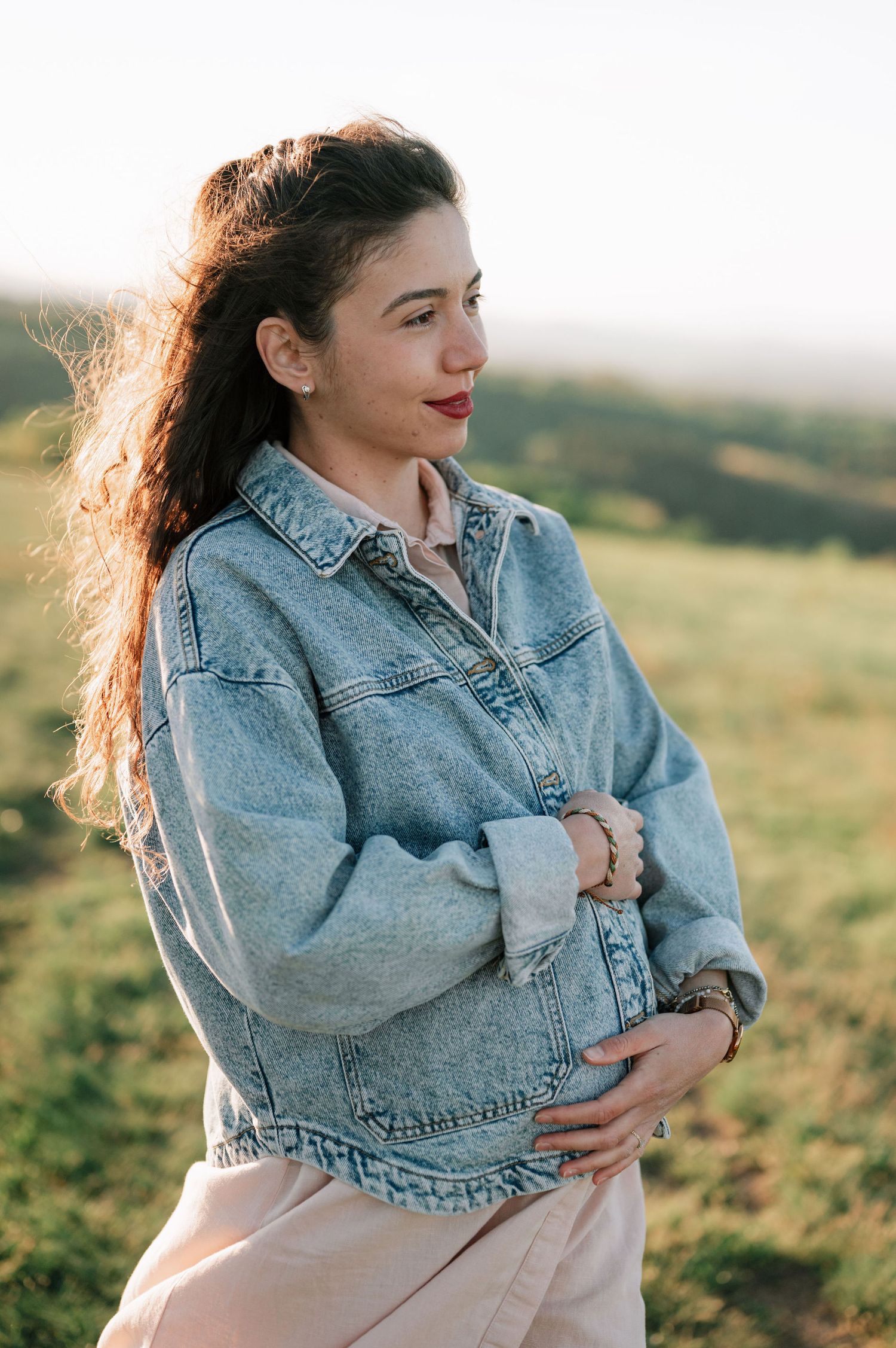 Woman in denim jacket poses thoughtfully in golden sunlit field at sunset, hair blowing in the breeze.
