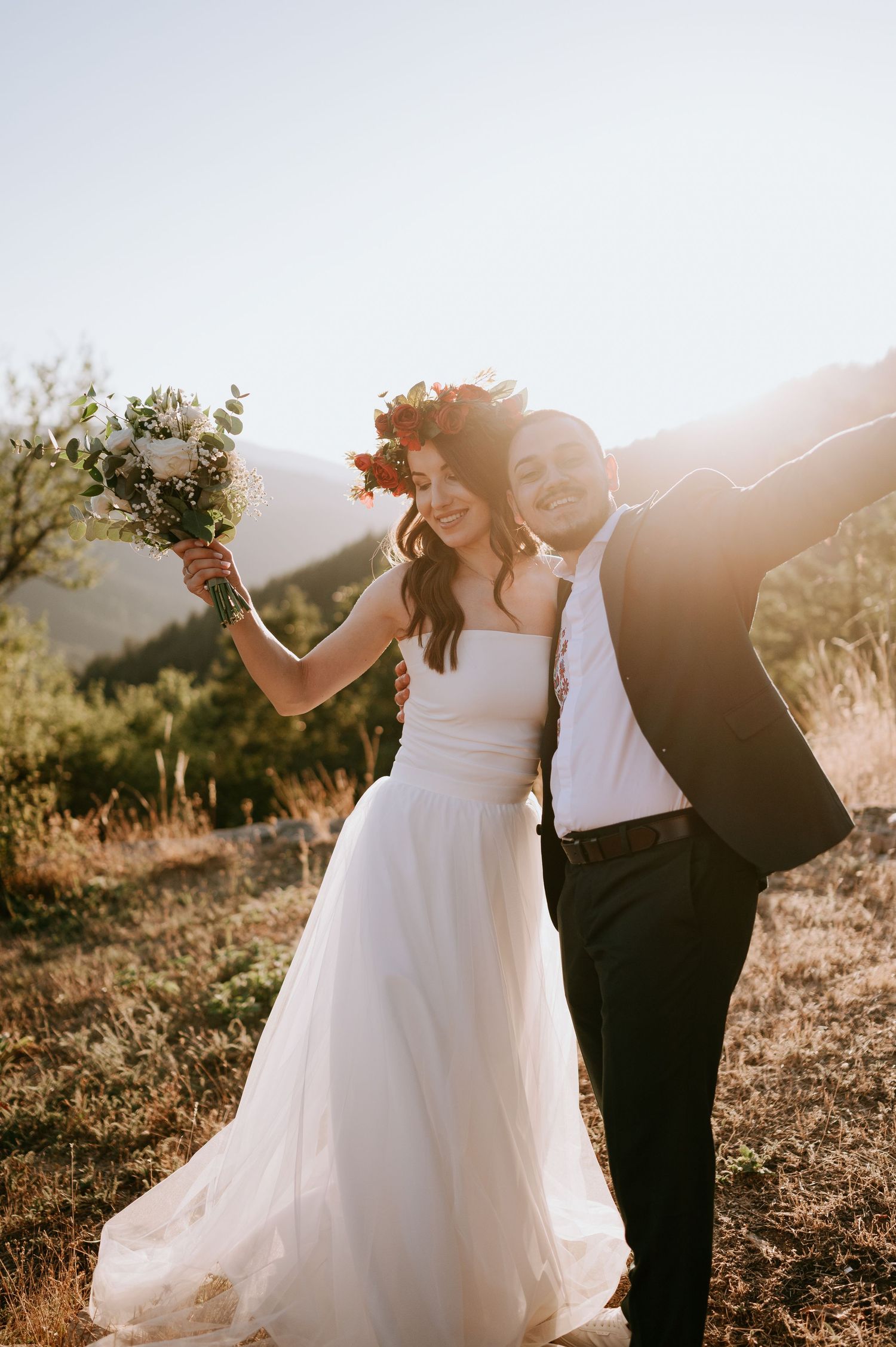 Wedding couple poses playfully with bouquet against mountain backdrop during sunset.