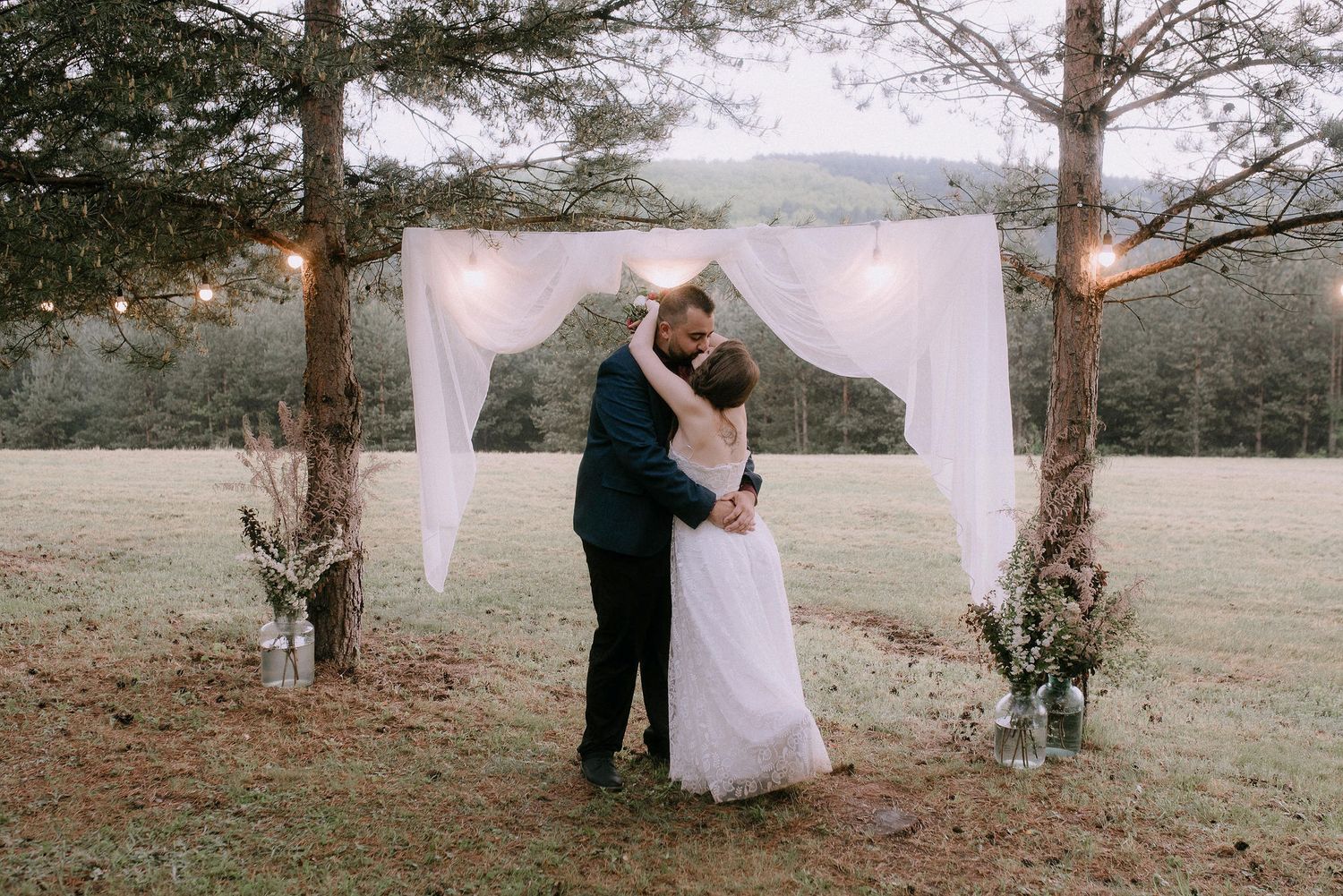 Wedding ceremony setup in natural setting with white fabric arch and pine trees.
