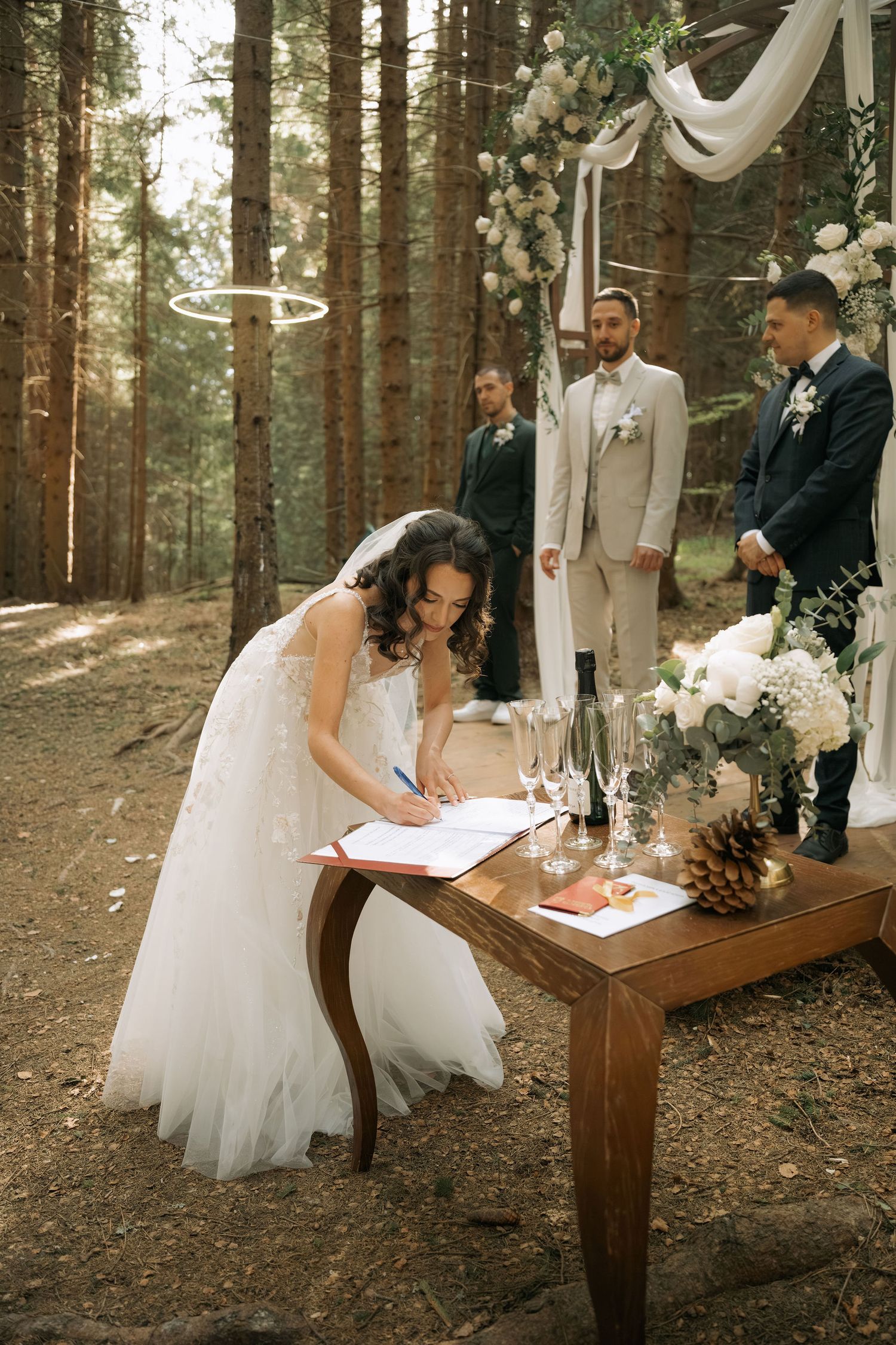Bride in white lace gown signing wedding register at wooden table in forest ceremony with groom and officiant.