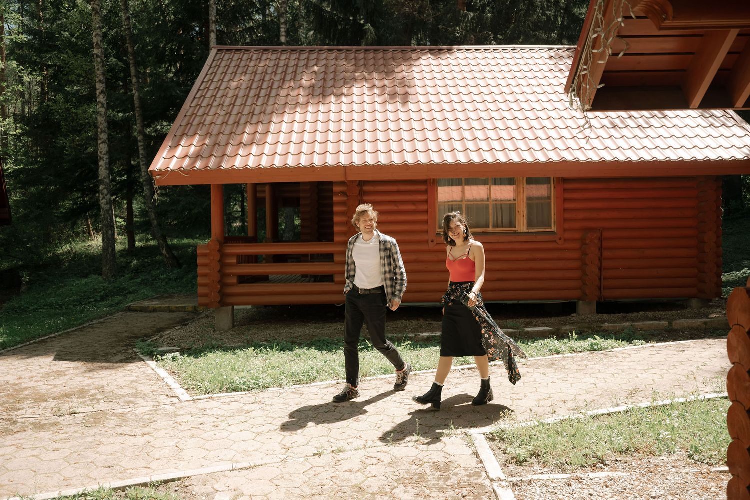 Couple walking hand in hand outside a rustic log cabin surrounded by tall pine trees.