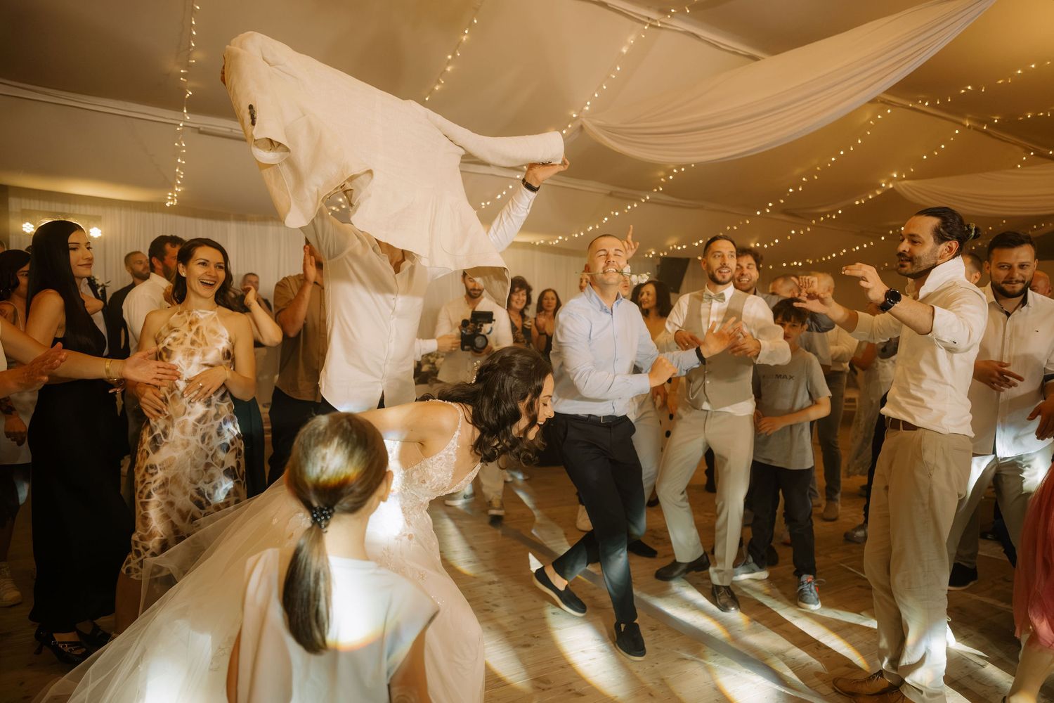 Joyful wedding reception dance floor with guests celebrating as bride and groom dance energetically under warm fairy lights.