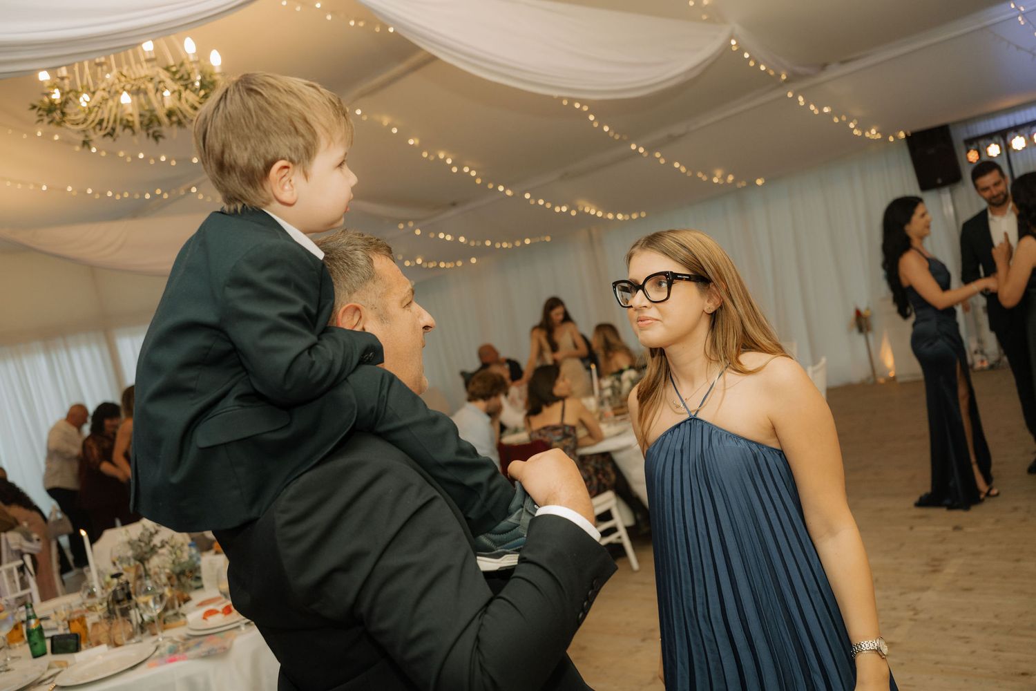 Man holding young boy in suit at elegant wedding reception while woman in blue dress and glasses looks on.