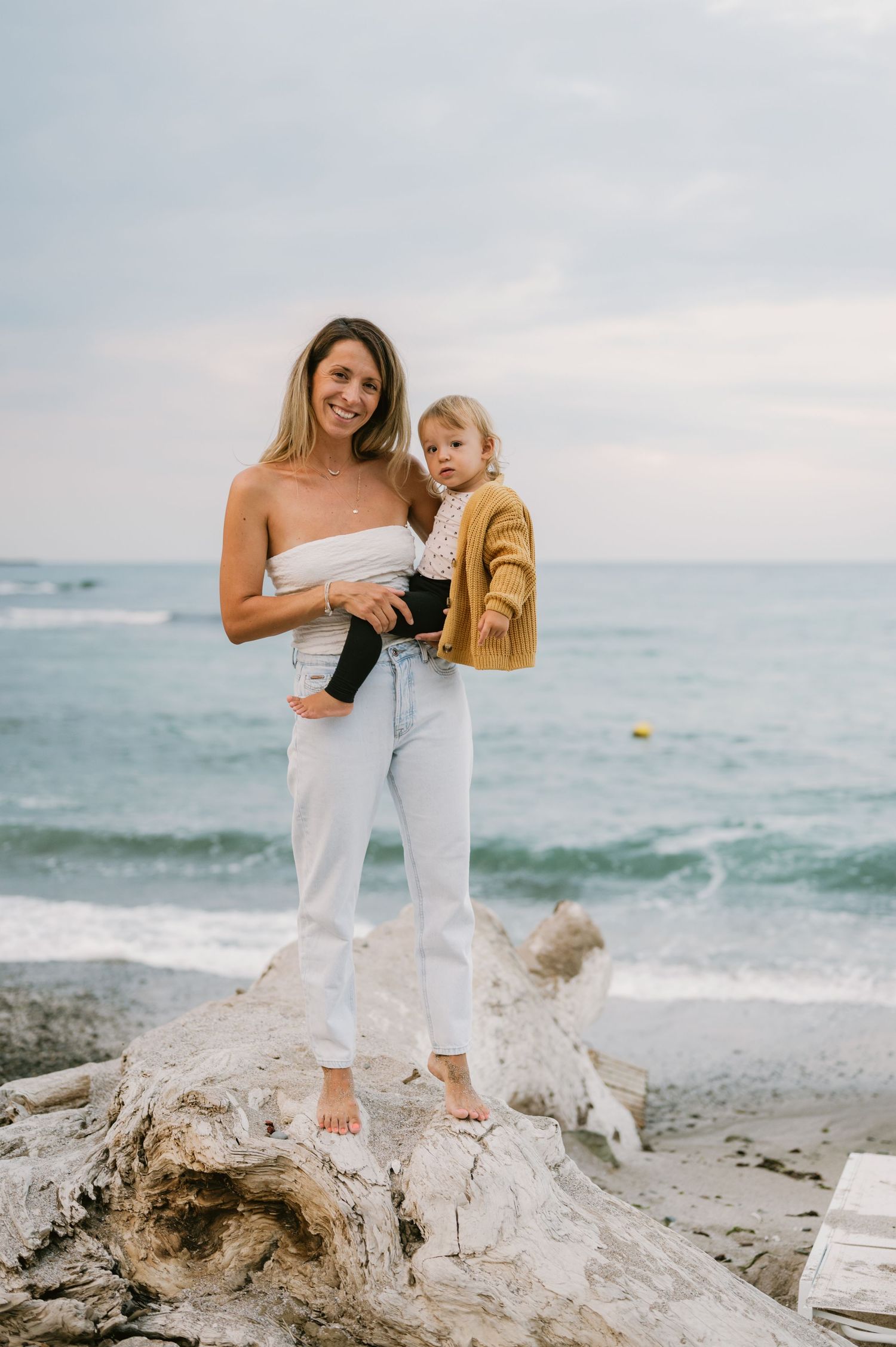 A smiling woman in white holds a toddler in a yellow cardigan while standing on driftwood at the beach.